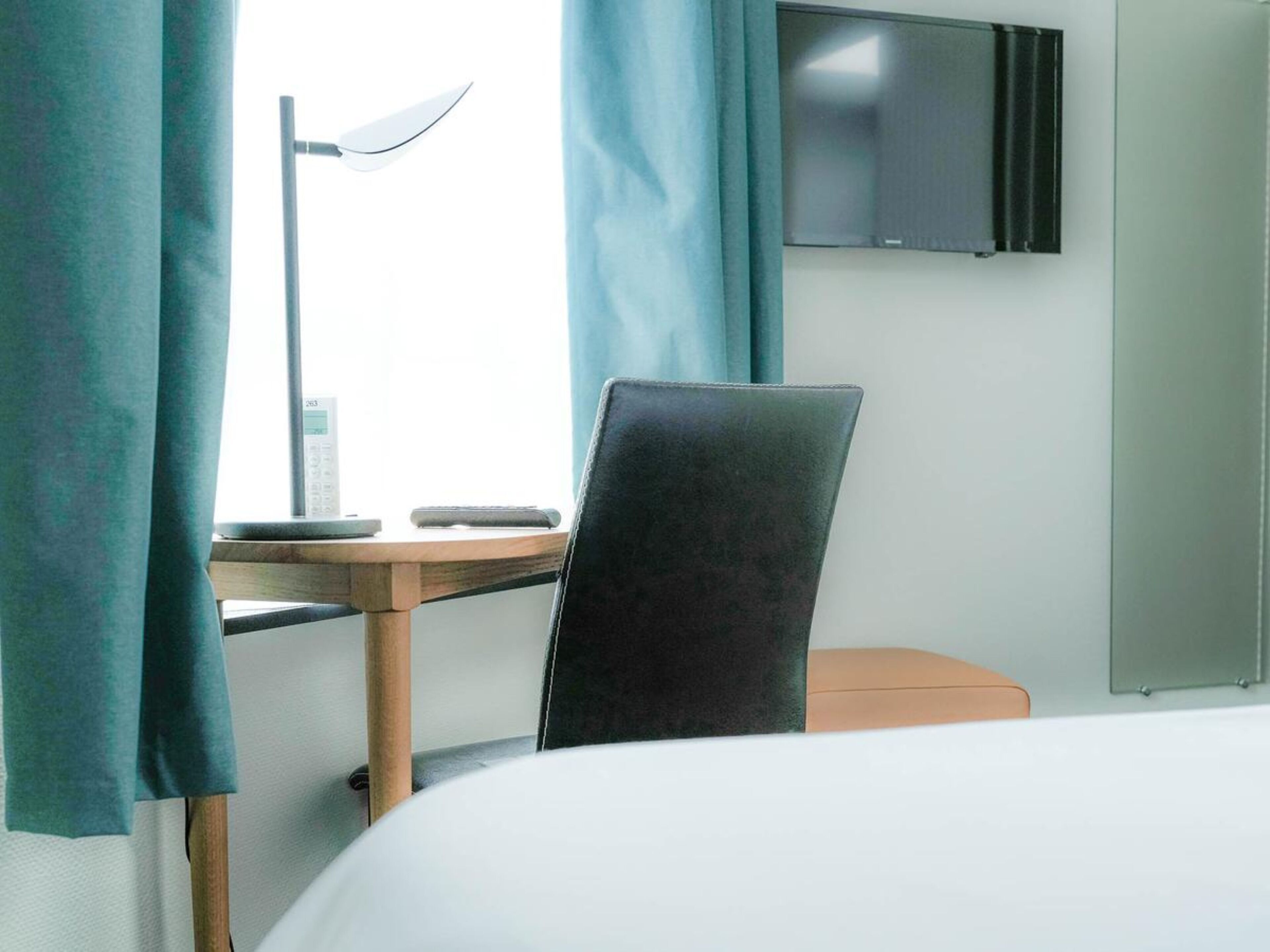 A hotel room with a bed, a desk with a lamp, a chair, a TV mounted on the wall, and a window with curtains.
