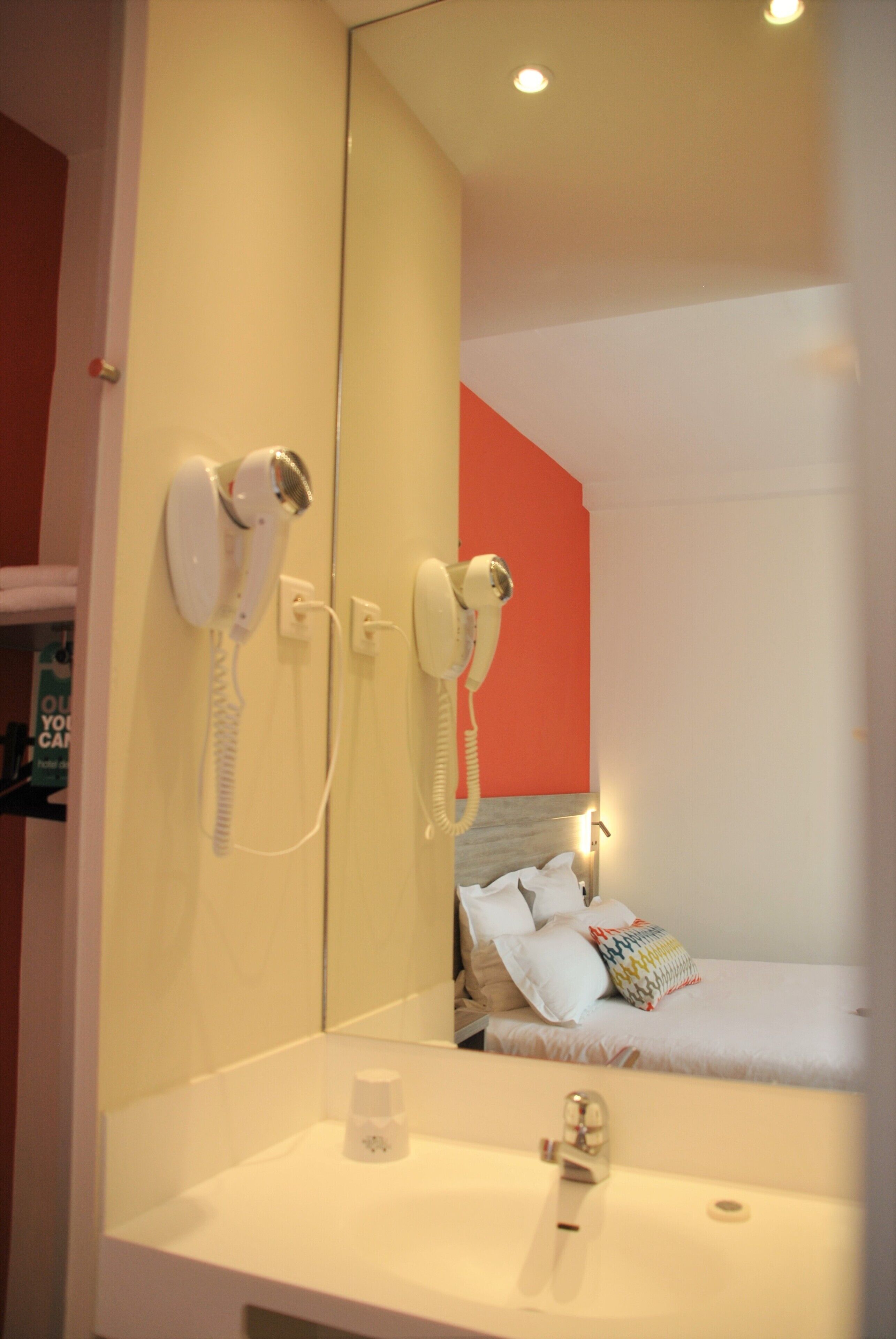 A hotel room with a large mirror reflecting a bed and a bathroom with two white hair dryers mounted on the wall.