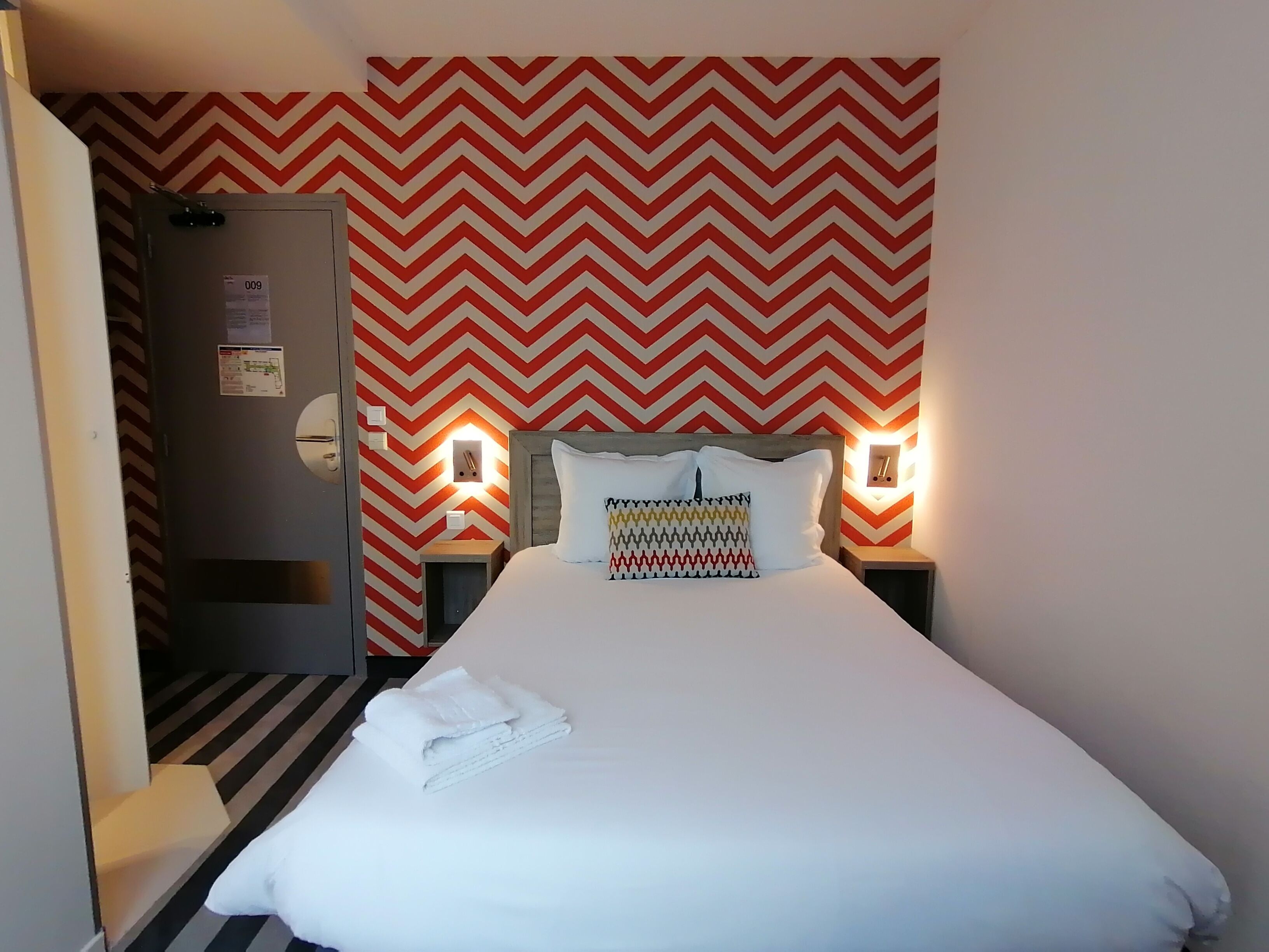 A hotel room with a bed, a nightstand, a wall clock, and a door with a number tag.