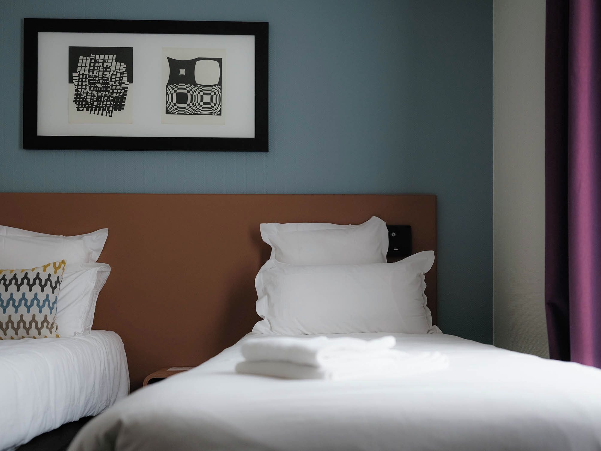 Habitación de hotel con dos camas, una obra enmarcada sobre ellas y una cortina púrpura a la derecha.
