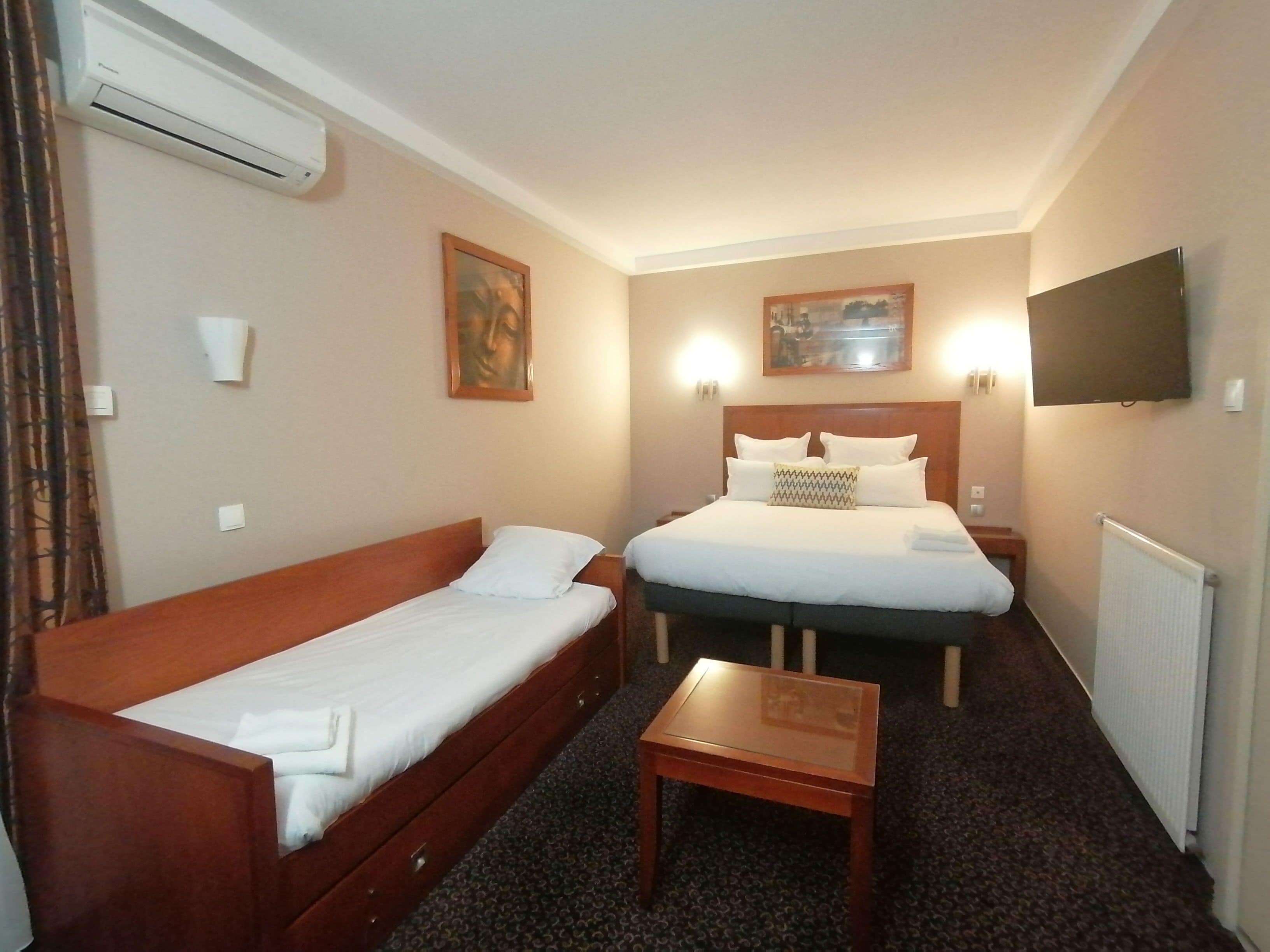 Una habitación de hotel con una cama individual, una cama doble, una mesita de noche de madera, una mesita pequeña y un equipo de aire acondicionado.