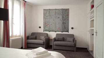 A hotel room with a bed, two armchairs, a radiator, a window with curtains, a wall-mounted artwork, and a wardrobe.