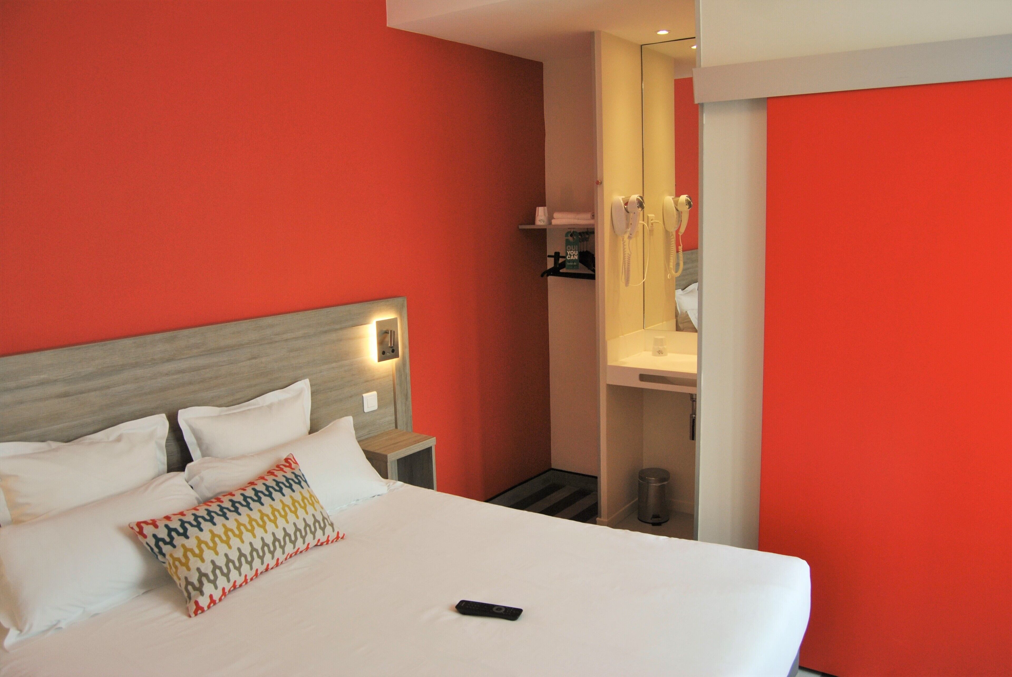 Habitación de hotel con cama, mesita de noche, espejo y pared roja.