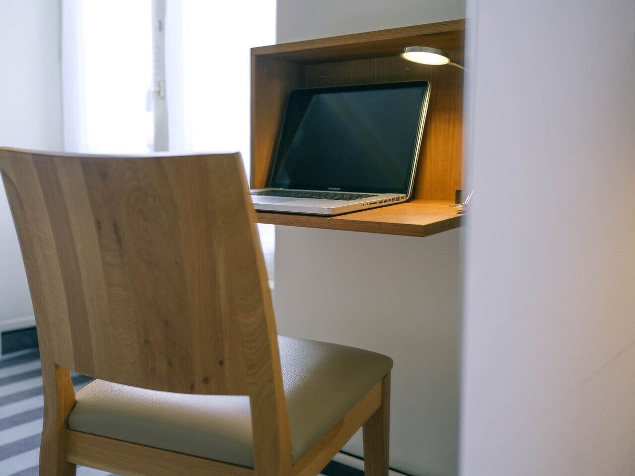 Una silla de madera con asiento y respaldo acolchados, ubicada frente a un soporte para laptop fijado a la pared, donde se encuentra una laptop.