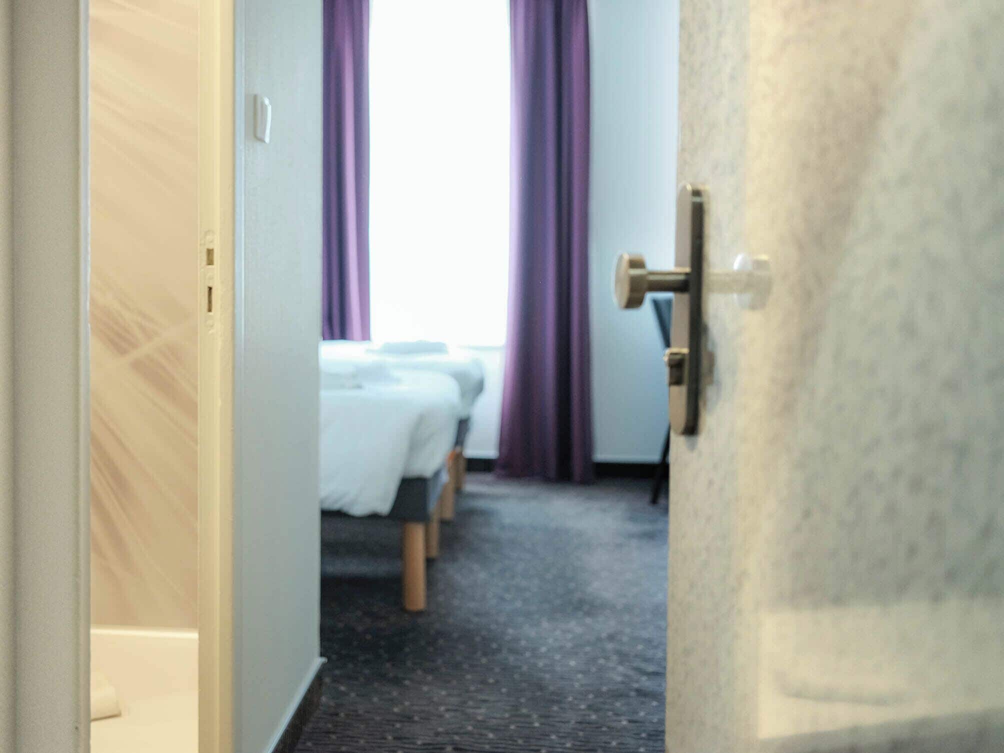 A hotel room with a bed, a desk, and a window with purple curtains.