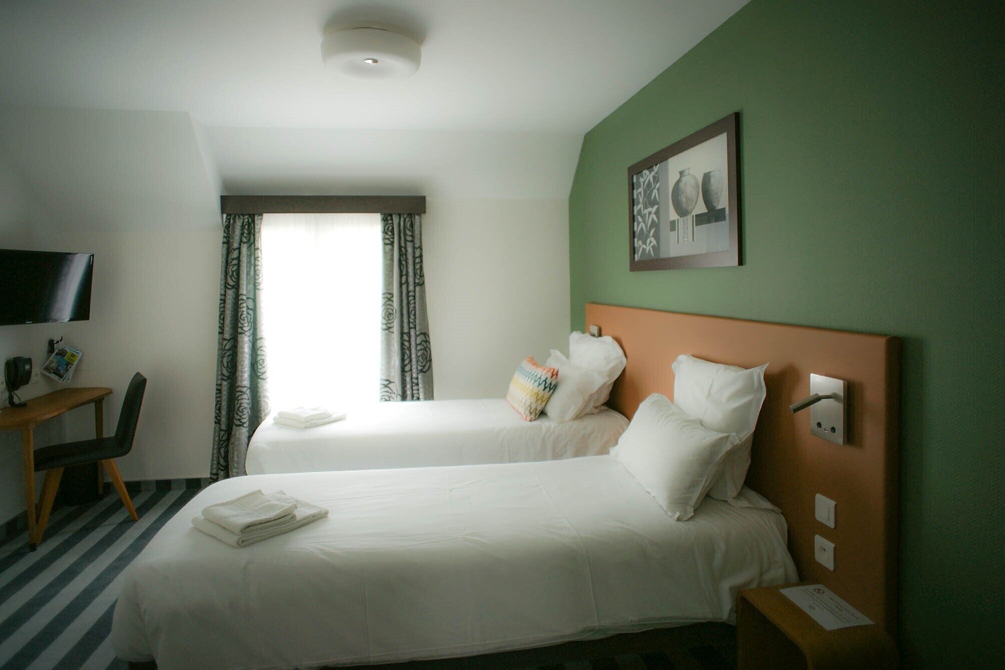 A hotel room with two beds, a desk, a TV, and a window with curtains.