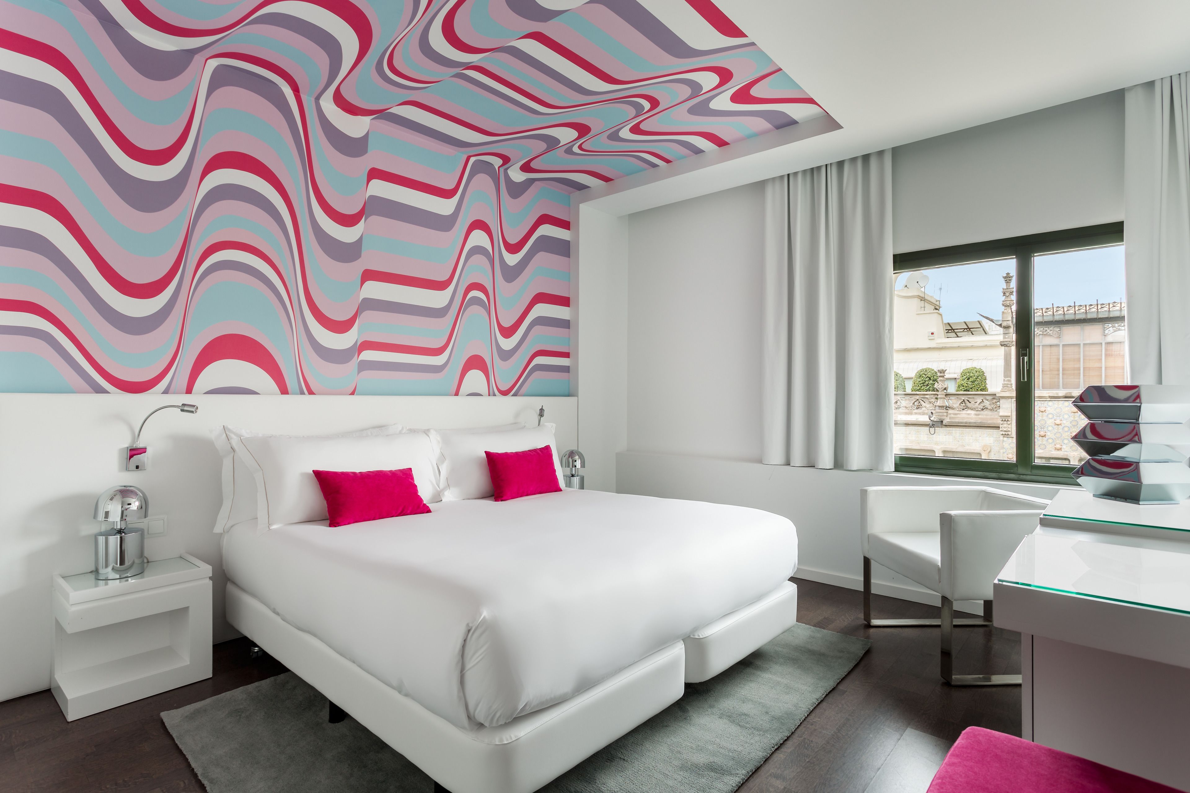 Habitación de hotel moderna con una cama grande, paredes blancas, un diseño ondulado y colorido en el techo, un escritorio y vistas al exterior.