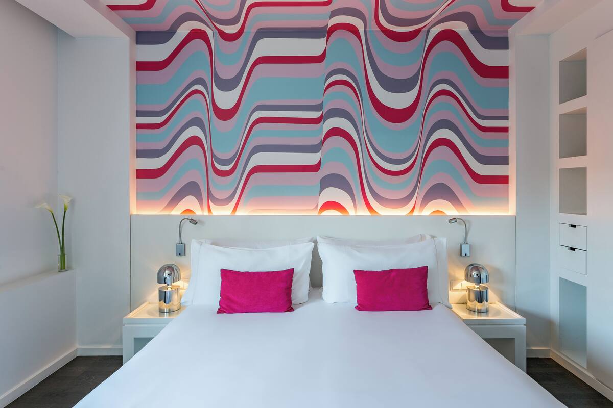 A modern hotel room with a large bed, two bedside tables with lamps, and a vibrant abstract wall mural.