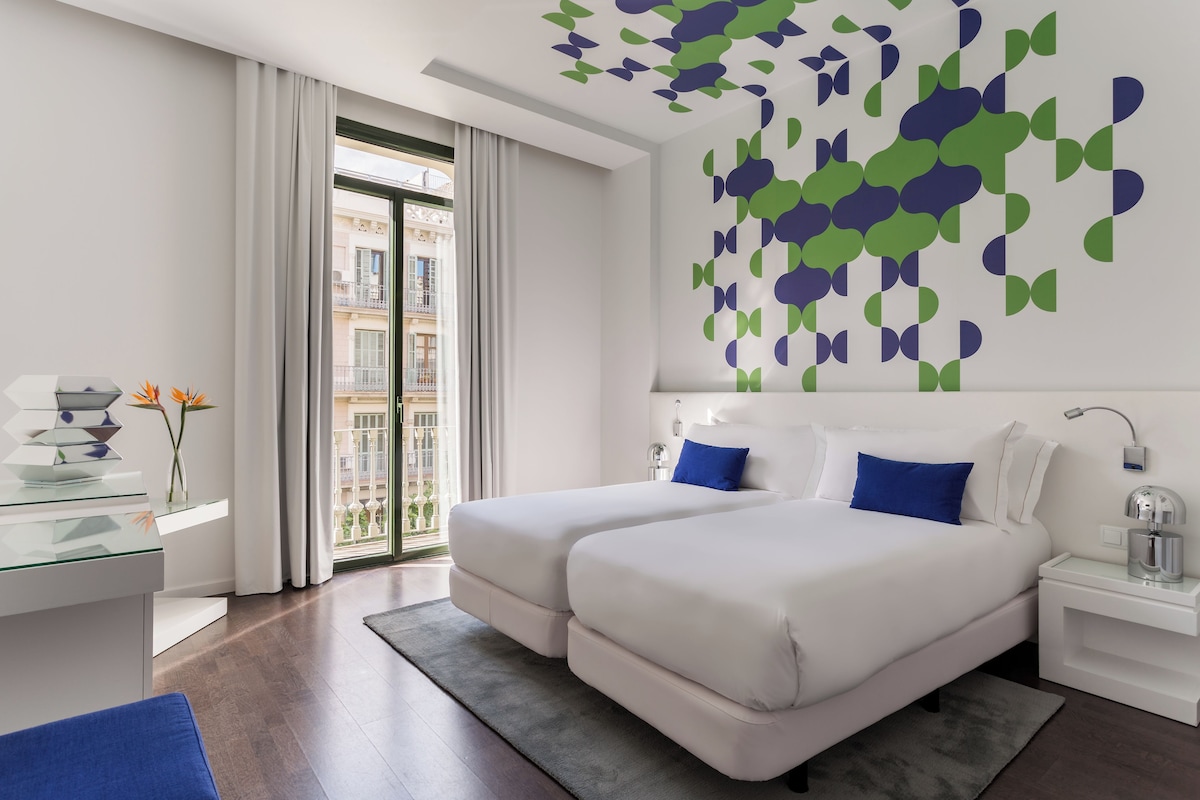 A modern hotel room with a balcony, a bed with blue pillows, a glass desk, and a decorative wall piece.