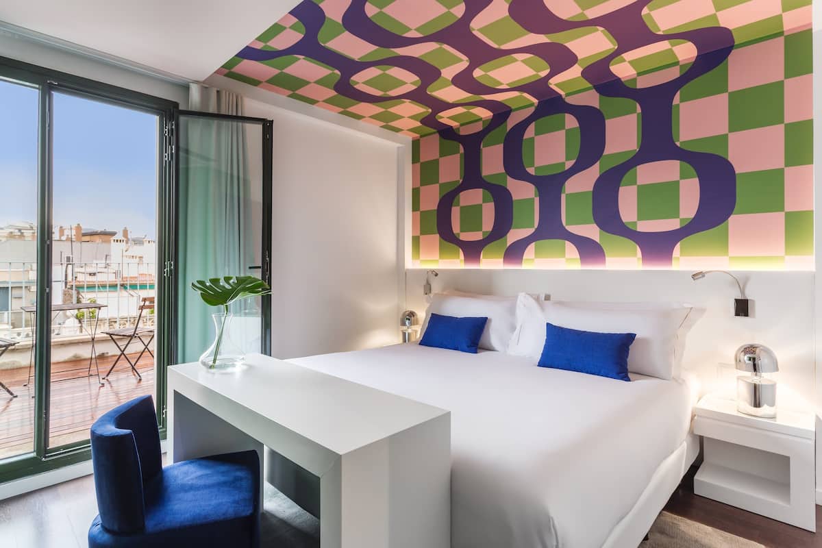 A modern hotel room with a large bed, a desk, and a view of the city through floor-to-ceiling windows.