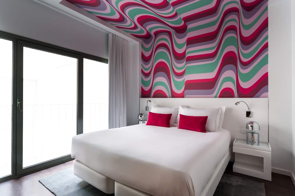 A modern hotel room with a large bed, white bedding, and a colorful abstract wall design.