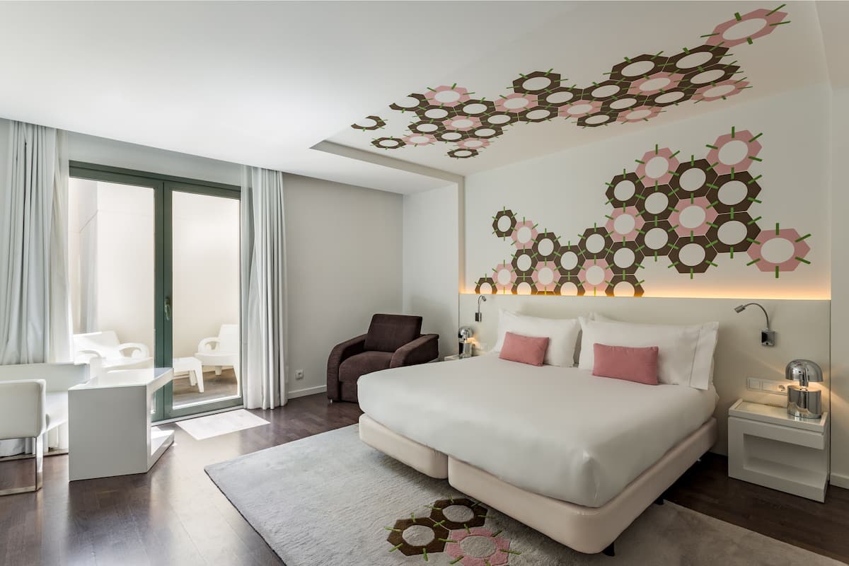 A modern hotel room with a large bed, a desk, a chair, and a view through a glass door.
