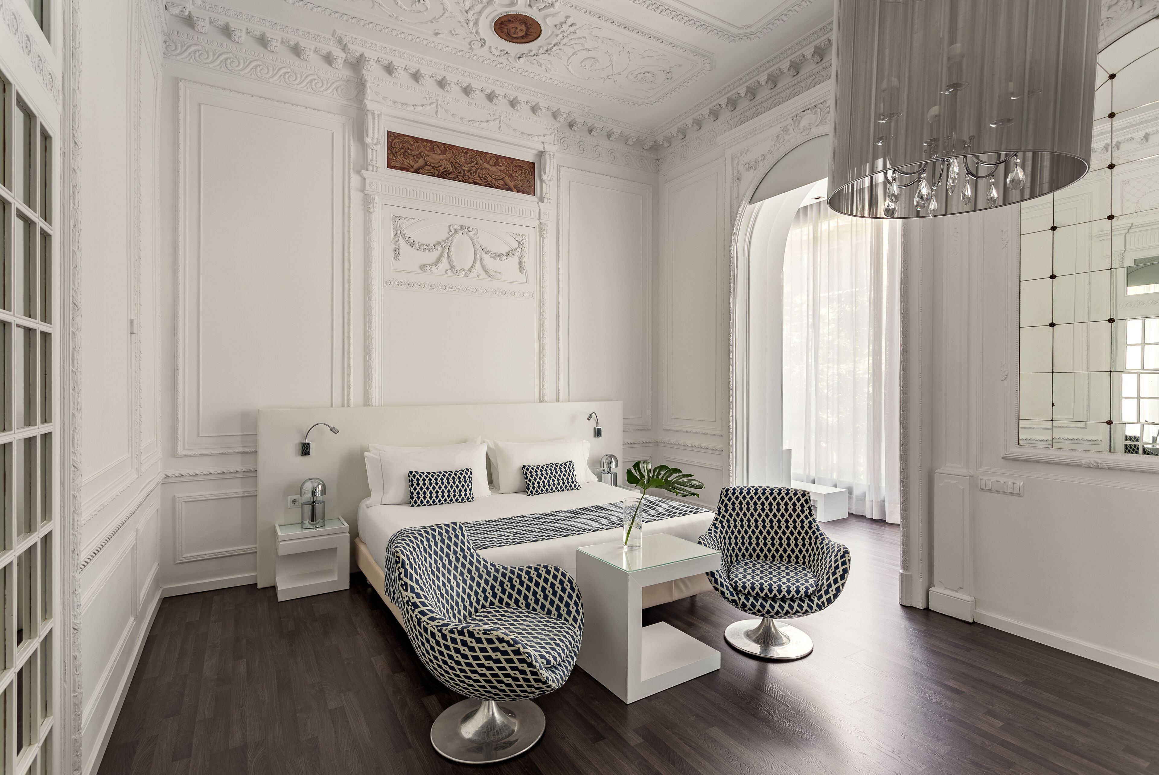 A modern bedroom with a large bed, two patterned chairs, a glass coffee table, and a chandelier.