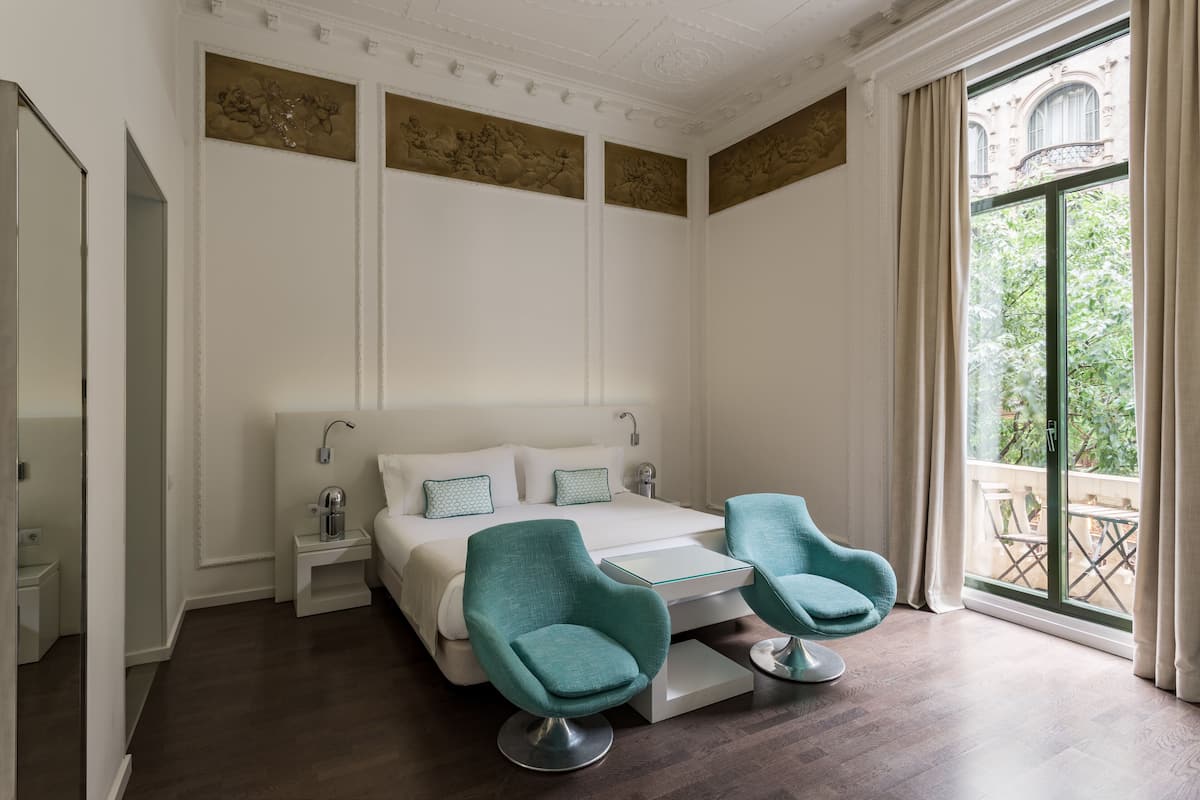 A modern hotel room with a large bed, two teal armchairs, a small table, and a balcony with a view of greenery.