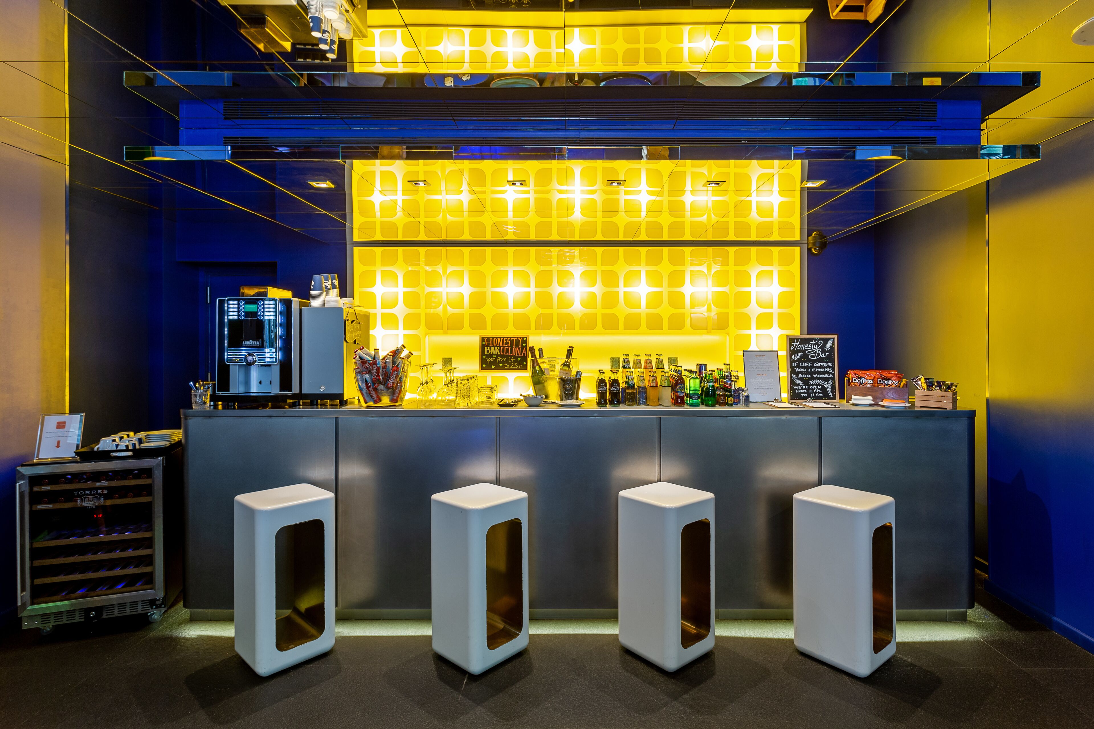 Een bar met een modern design, met een toonbank met een digitaal scherm, een selectie drankjes en drie witte barkrukken.