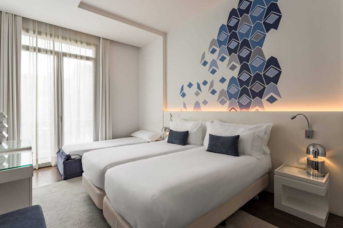 A hotel room with two beds, a glass desk, a wall-mounted coffee machine, and a geometric wall decoration.