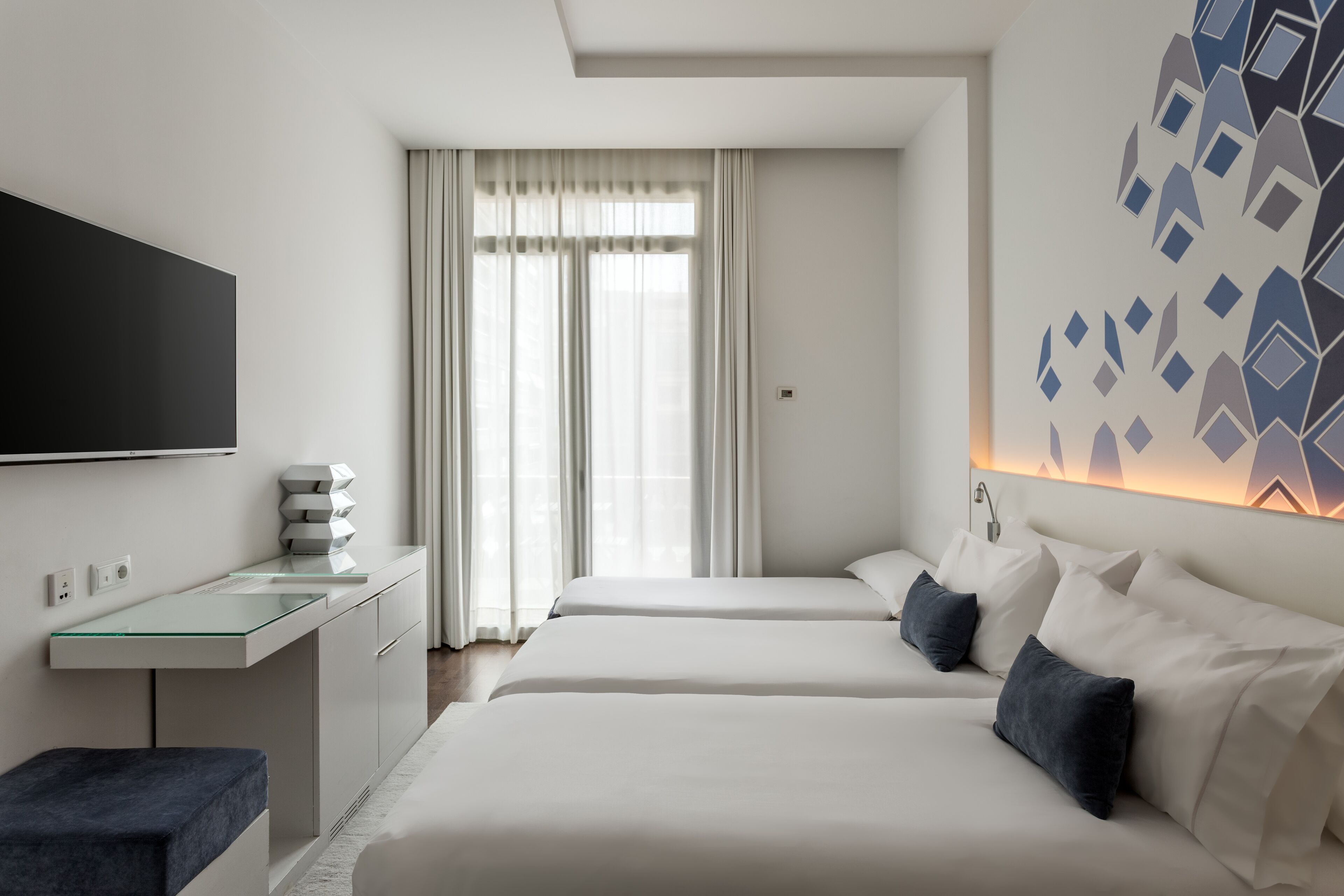 Habitación de hotel moderna con dos camas, un escritorio de cristal, un televisor de pantalla plana y un diseño geométrico en la pared.