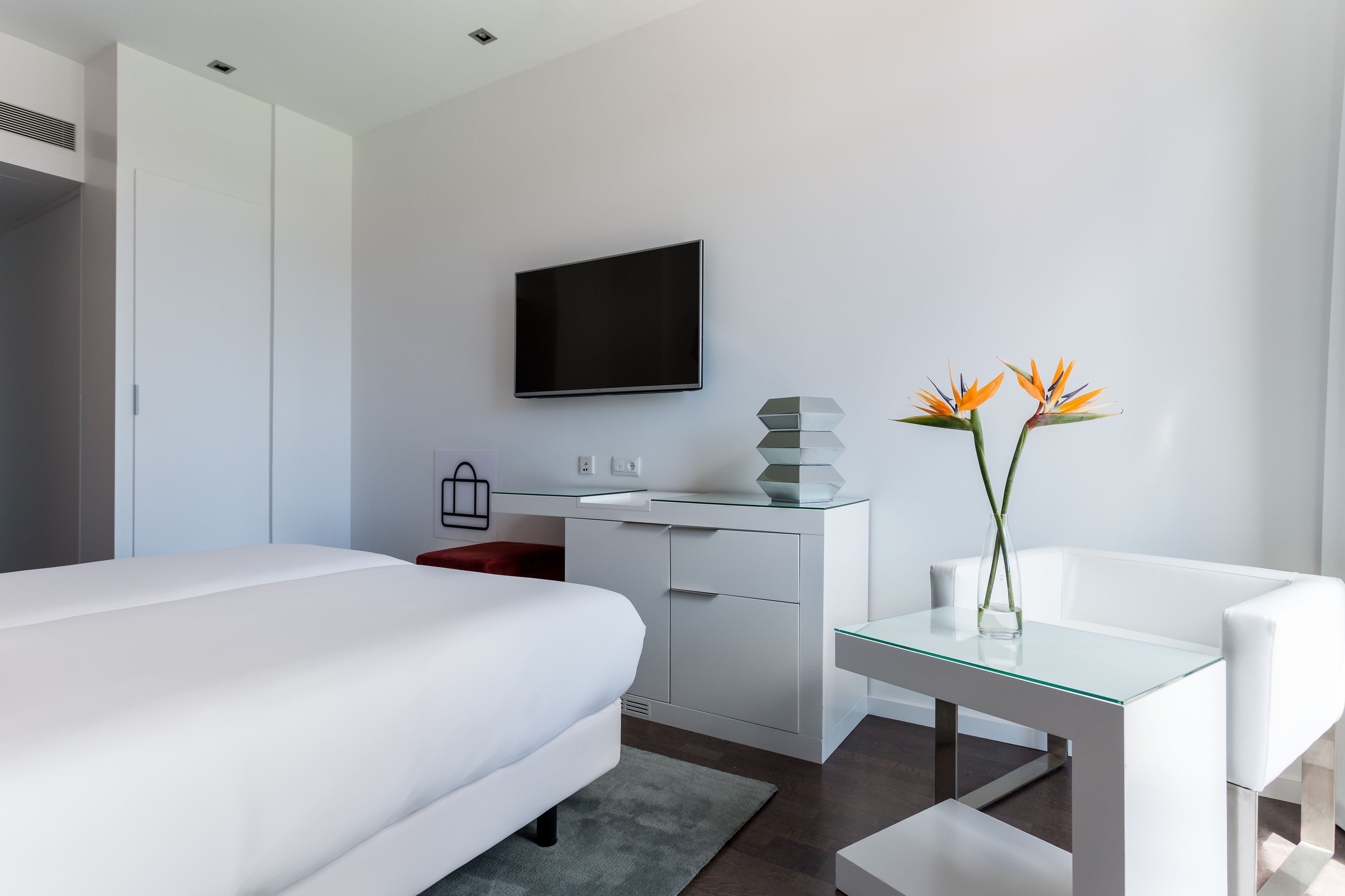 A modern hotel room with a large bed, a desk with a flat-screen TV, a vase with flowers, and a white chair.