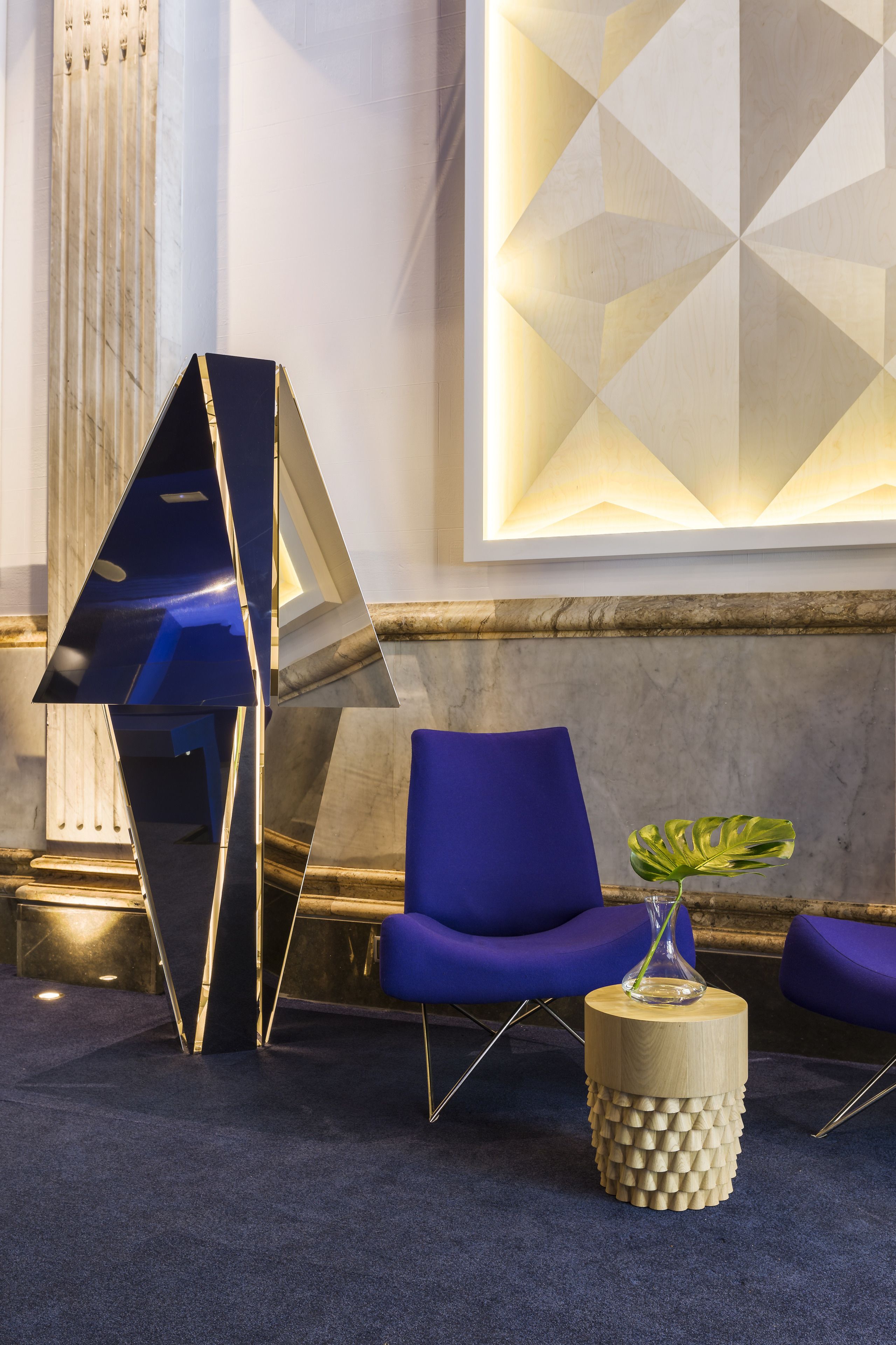 A modern lobby with a blue chair, a unique geometric sculpture, and a round wooden side table with a plant.