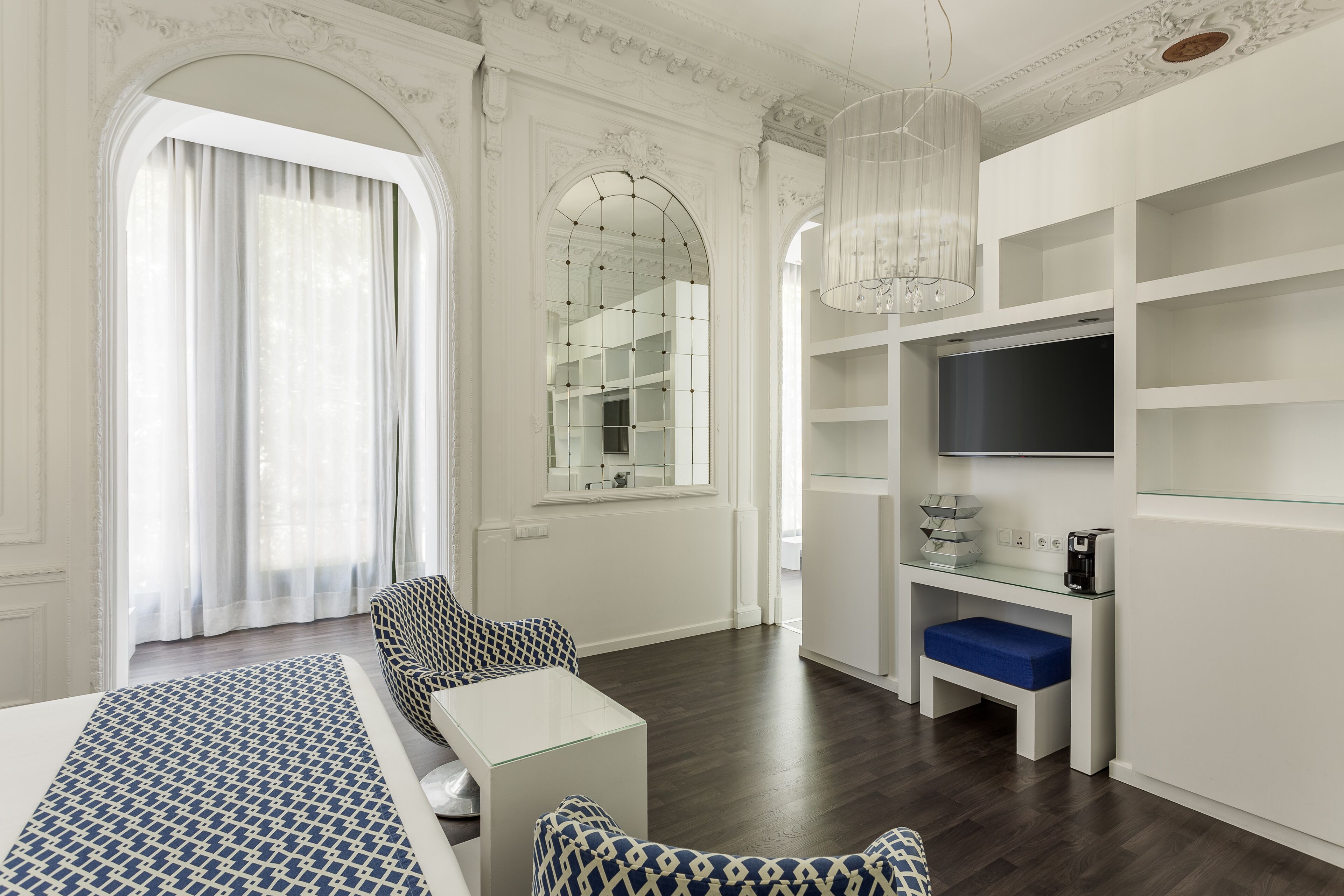 Een moderne hotelkamer met een groot bed, een zithoek met gestreepe banken, een televisie op een witte standaard en een groot, rondbogig raam met gordijnen.