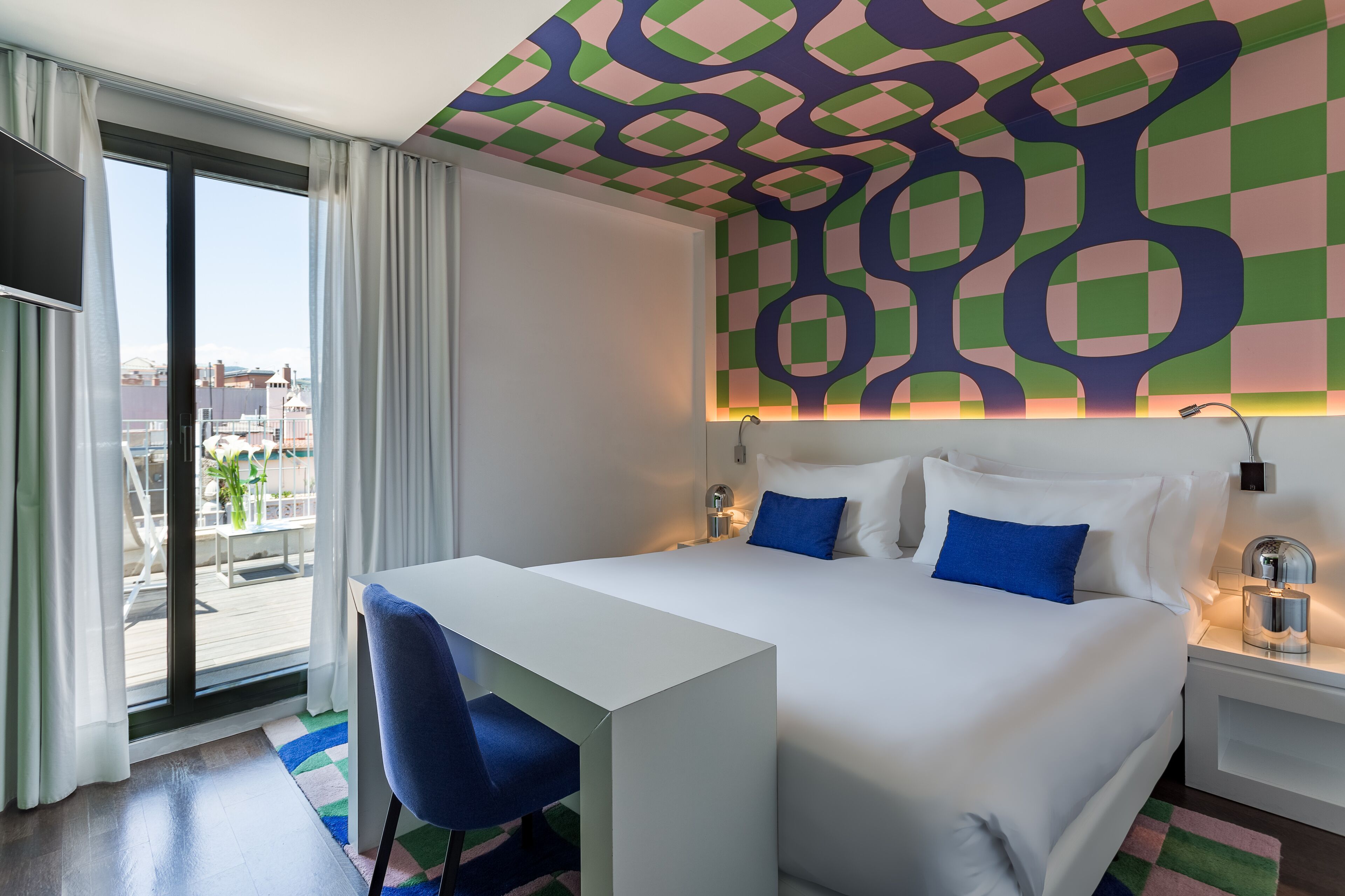 A hotel room with a large bed, a desk, and a view of the city through a sliding door.