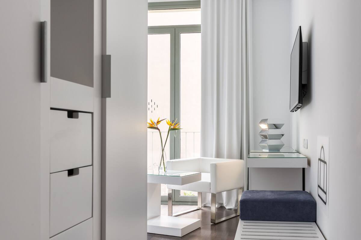 A modern hotel room with a minimalist design, featuring a white color scheme, a seating area with a bench, a small table, and a television mounted on the wall.