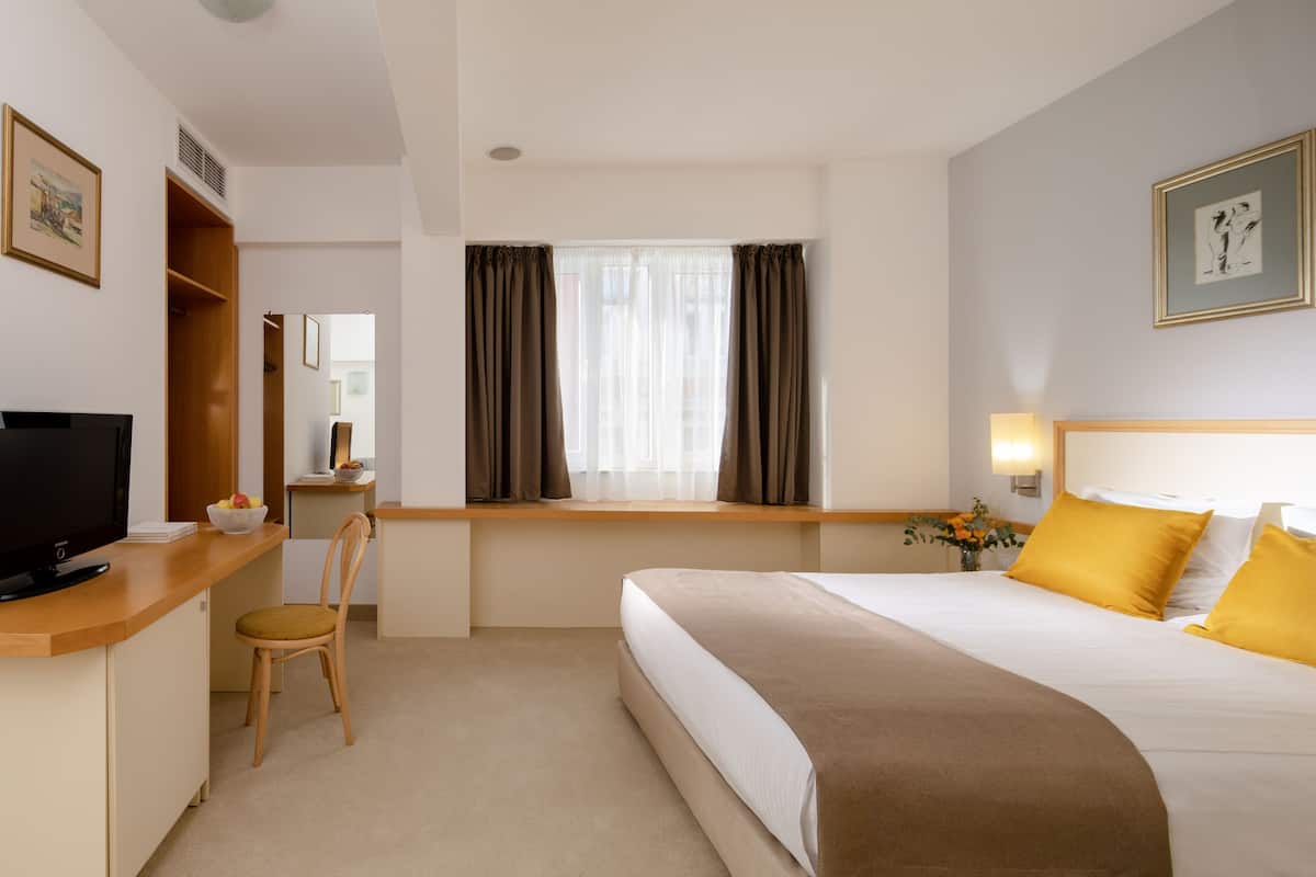 A hotel room with a bed, a desk with a television, a chair, a window with curtains, and a bedside table with a lamp.