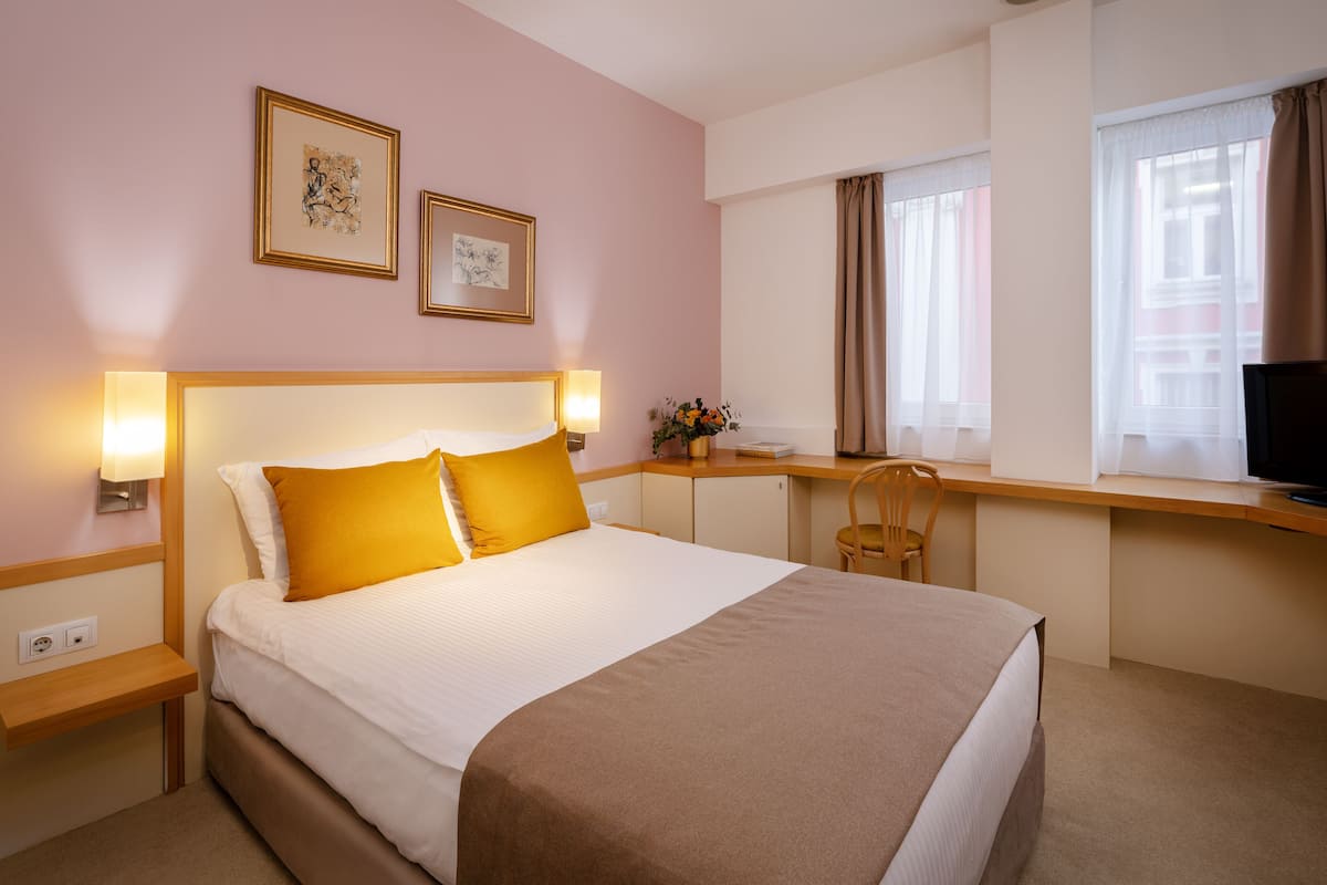 A hotel room with a bed, bedside lamps, a desk, a chair, and a TV.