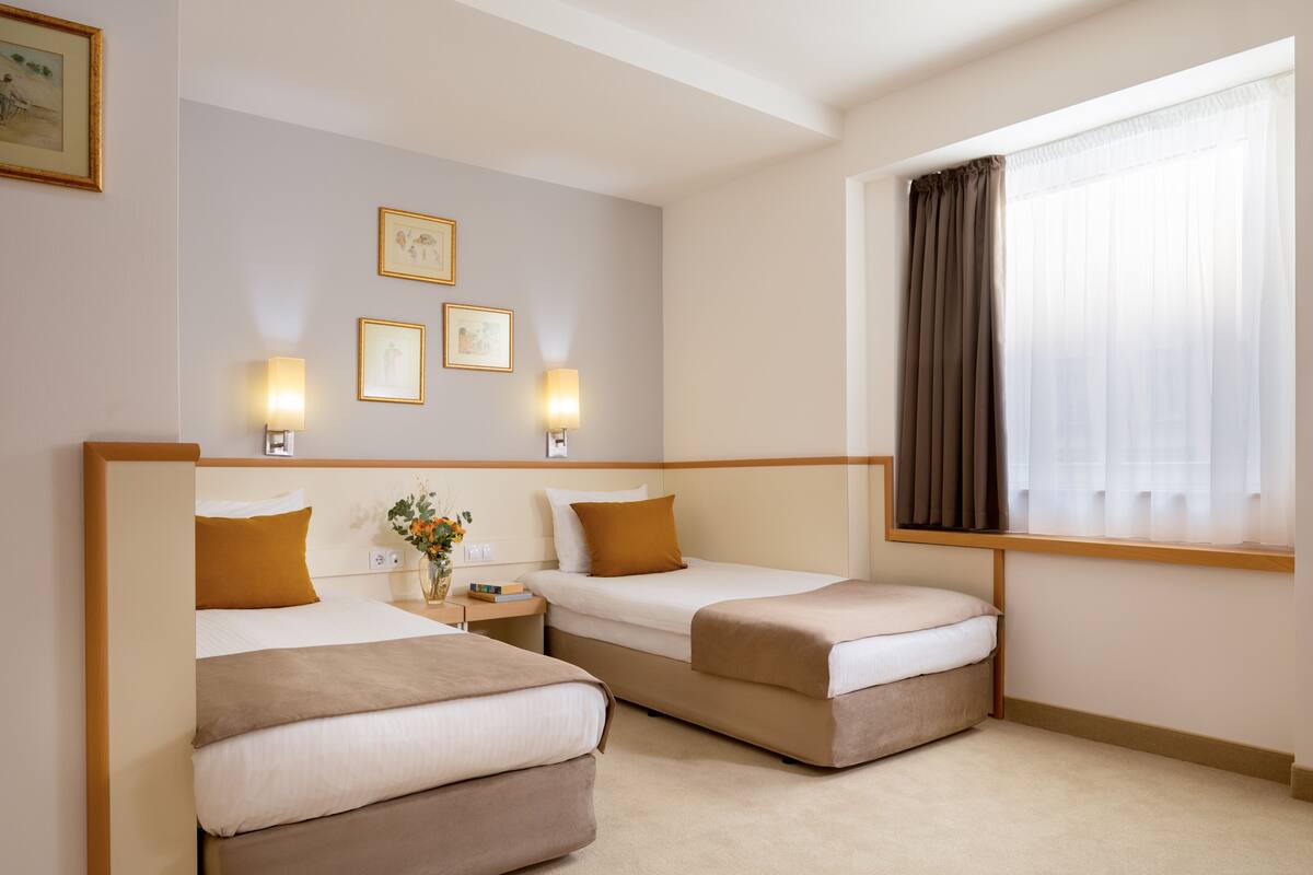 A hotel room with two beds, a nightstand with a lamp, framed pictures on the wall, and a window with curtains.