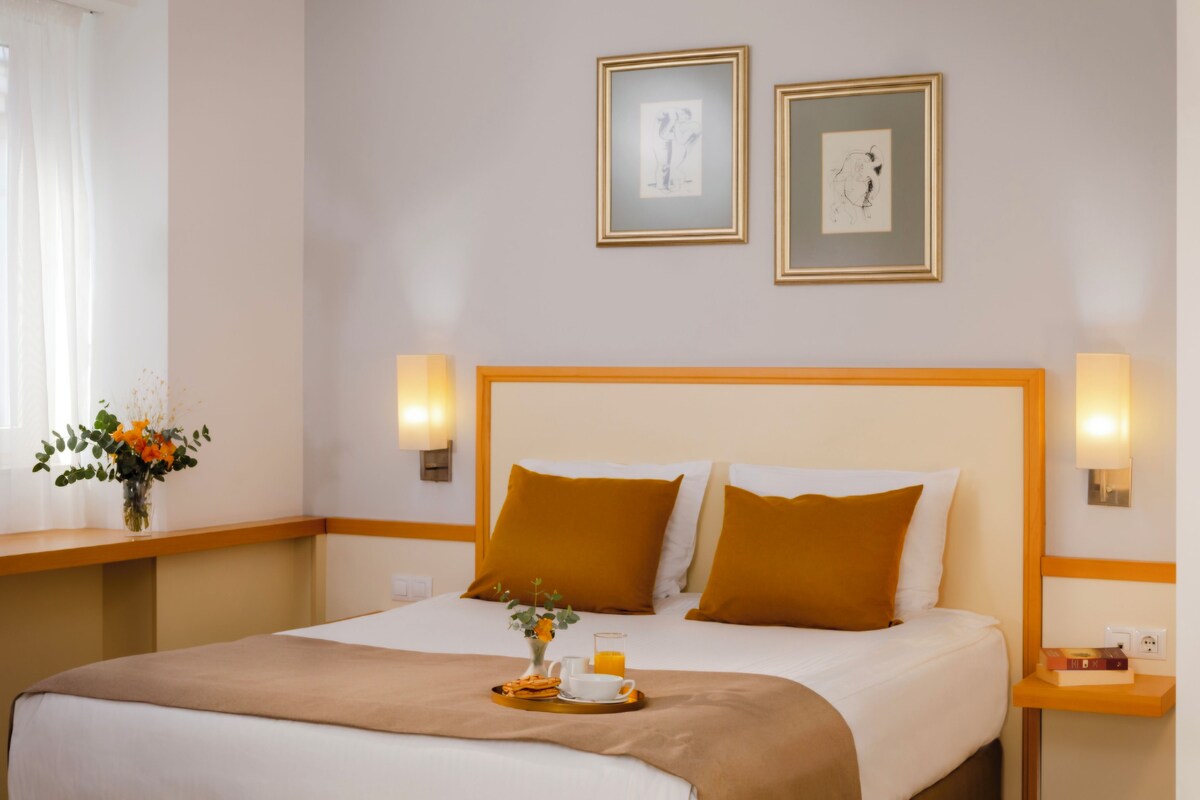 A hotel room with a bed, two wall-mounted lamps, a bedside table with a book, and a window with curtains.