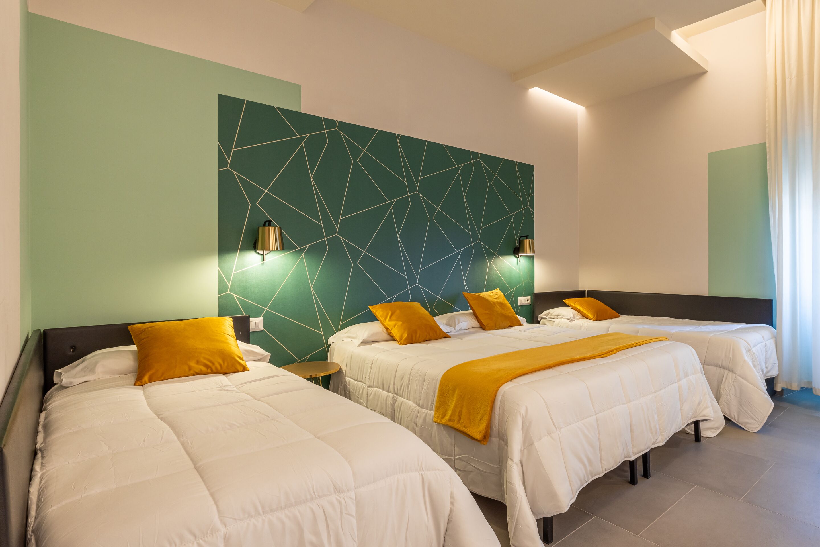 A hotel room with three beds, each with white bedding and a mustard-colored throw blanket. The room features a geometric-patterned accent wall and two wall-mounted lamps.