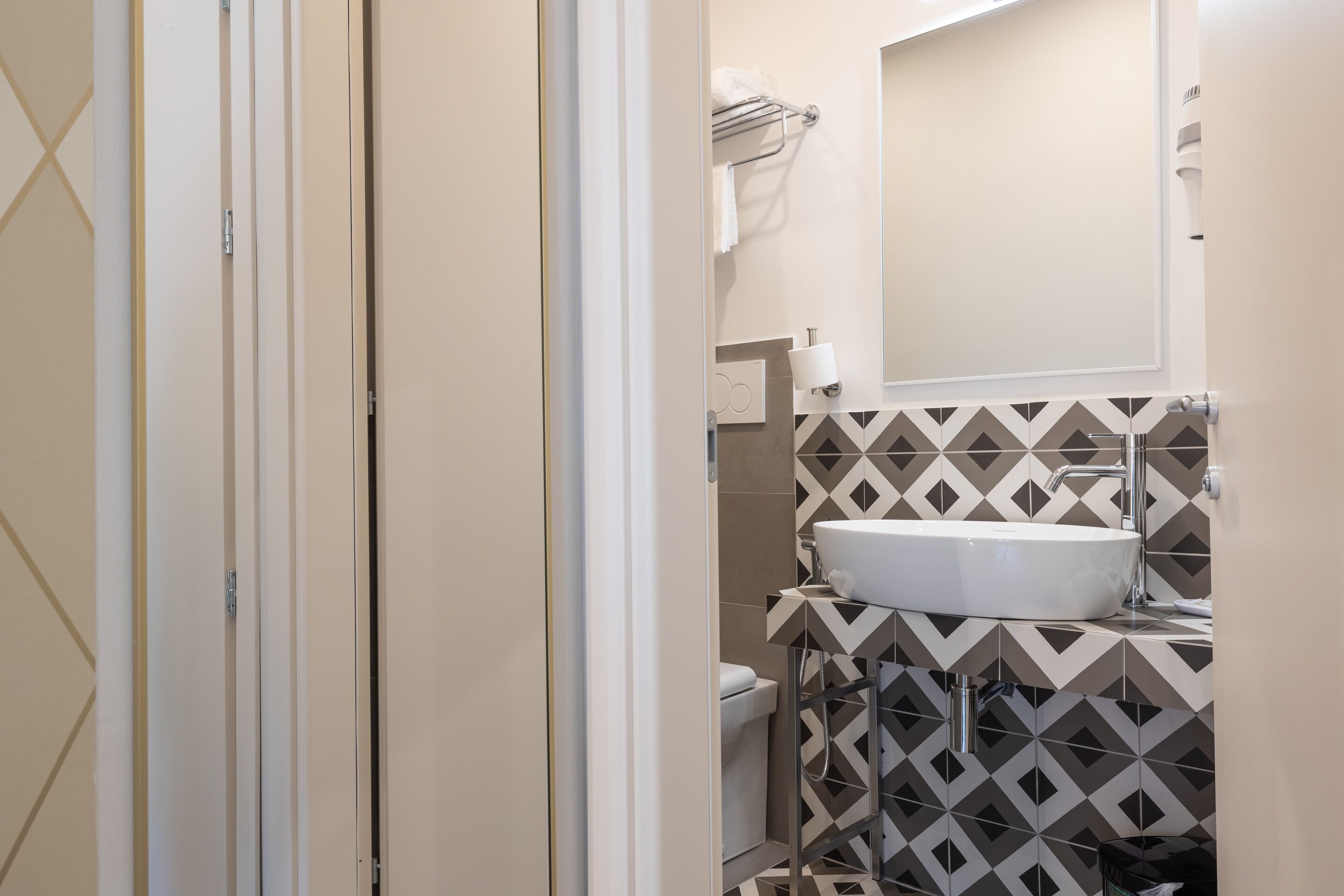 Une salle de bain avec un lavabo blanc, des toilettes et un miroir au-dessus du lavabo. Les murs sont recouverts de carreaux disposés selon un motif géométrique.