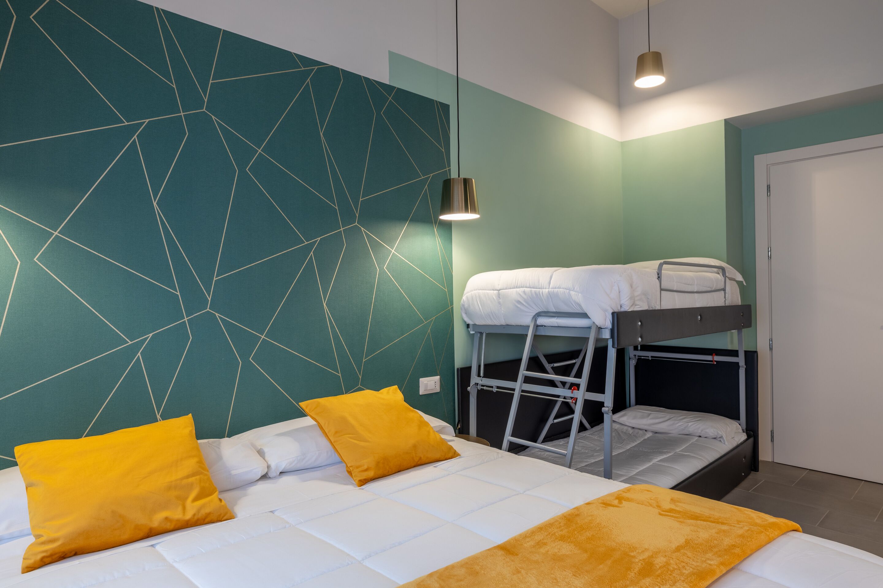 Ein Zimmer mit einem Etagenbett, einem Schreibtisch und einem Stuhl. Es gibt ein Fenster mit Vorhängen, eine Wand mit geometrischem Muster und eine Pendelleuchte.