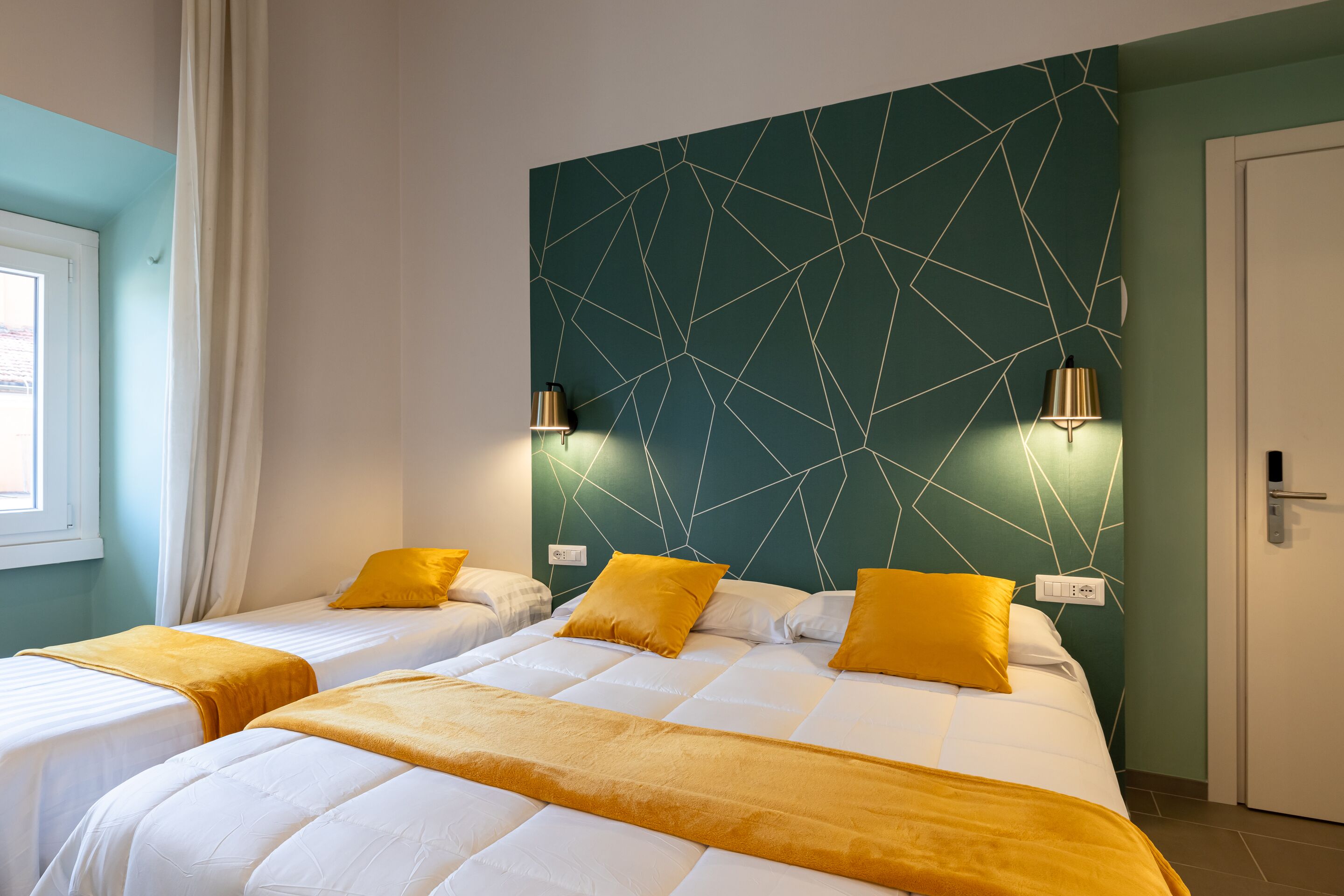 Une chambre d’hôtel avec deux lits, chacun agrémenté de draps blancs et de coussins décoratifs jaunes. La chambre est dotée d’une tête de lit à motifs géométriques et de luminaires fixés au mur.