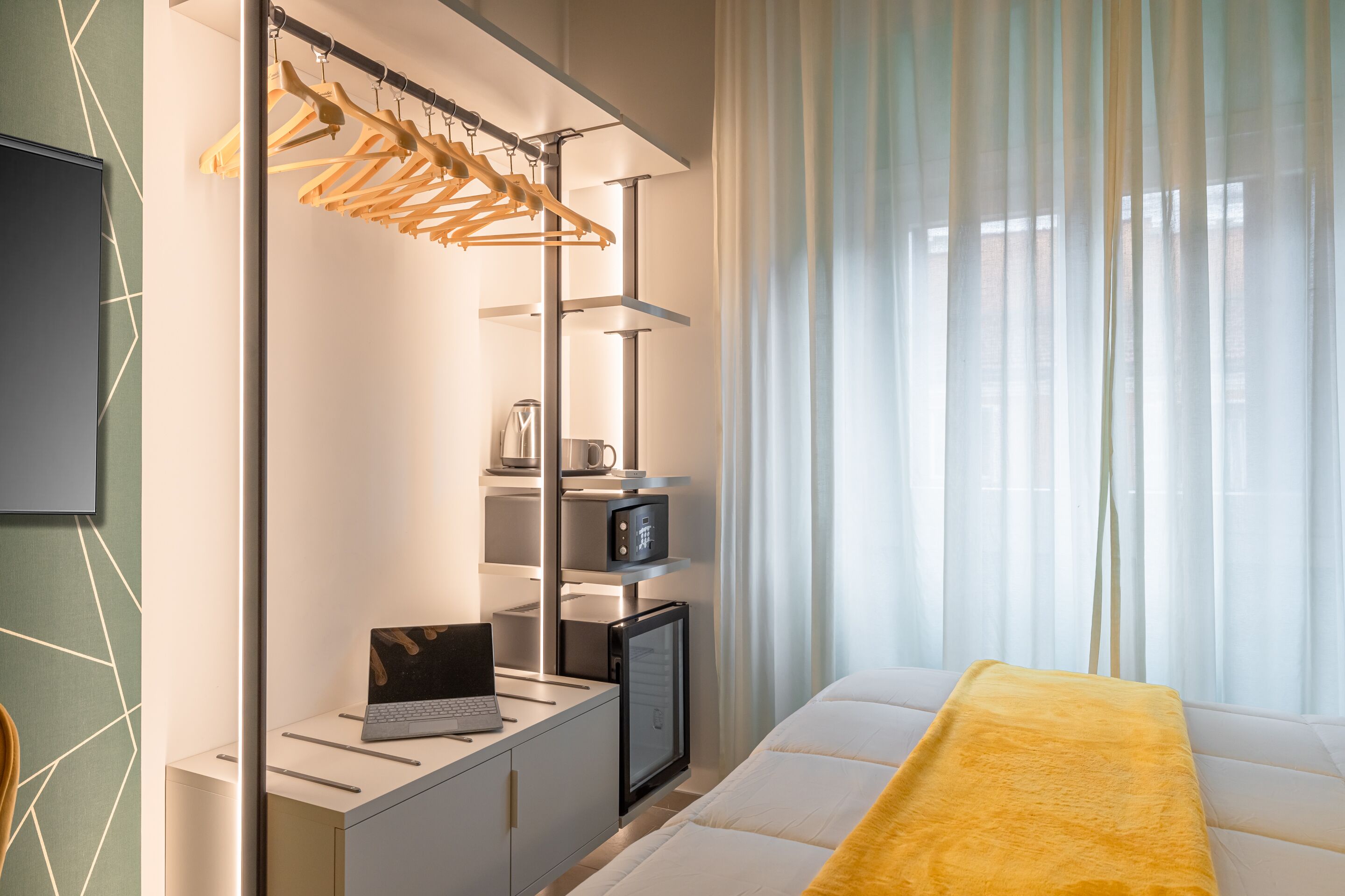 A hotel room with a bed, a desk with a laptop, a mini-fridge, a coffee maker, and a hanging clothes rack.