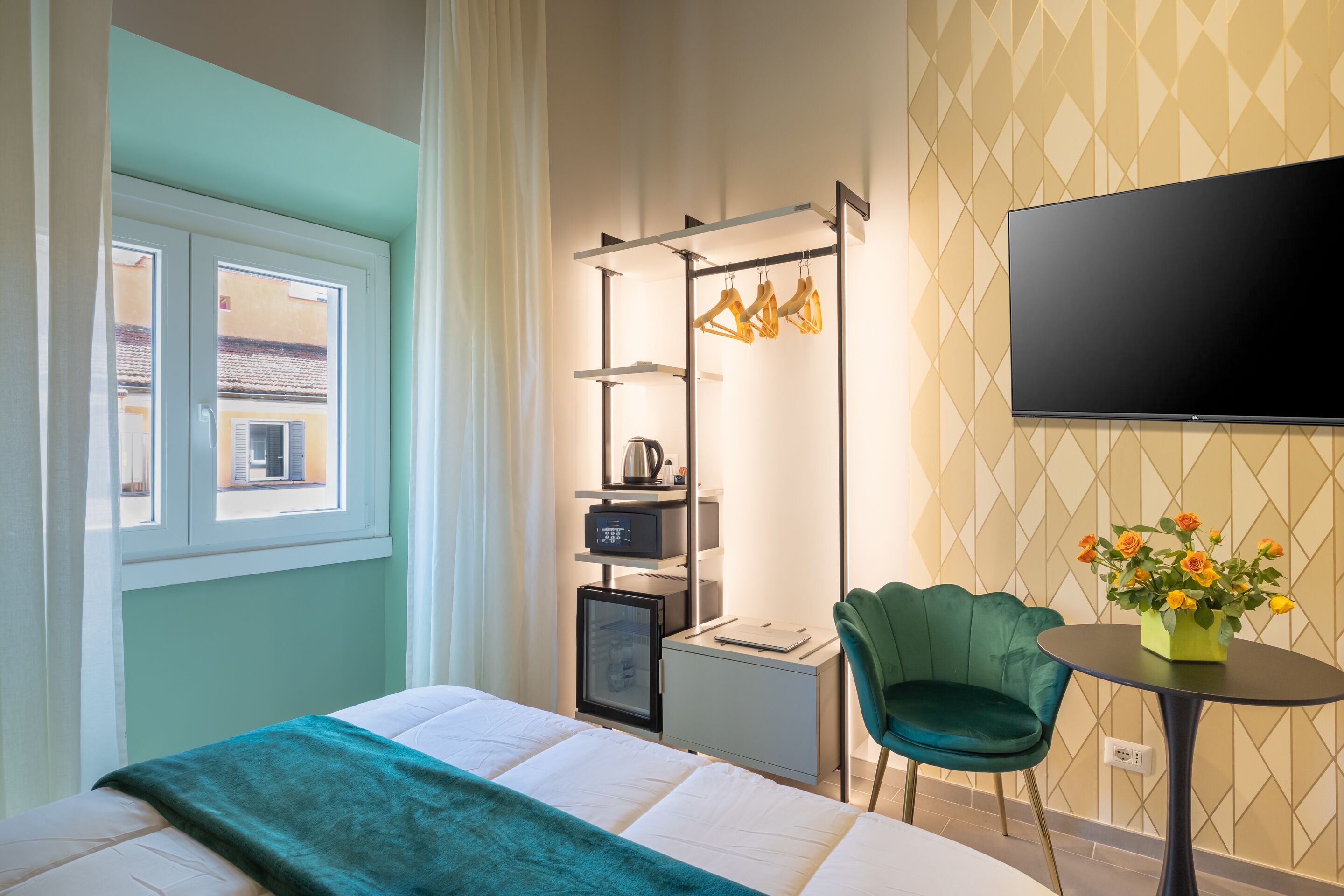 Una camera d'albergo con un letto, una scrivania con un computer, una sedia, una TV e una finestra vista edifici.