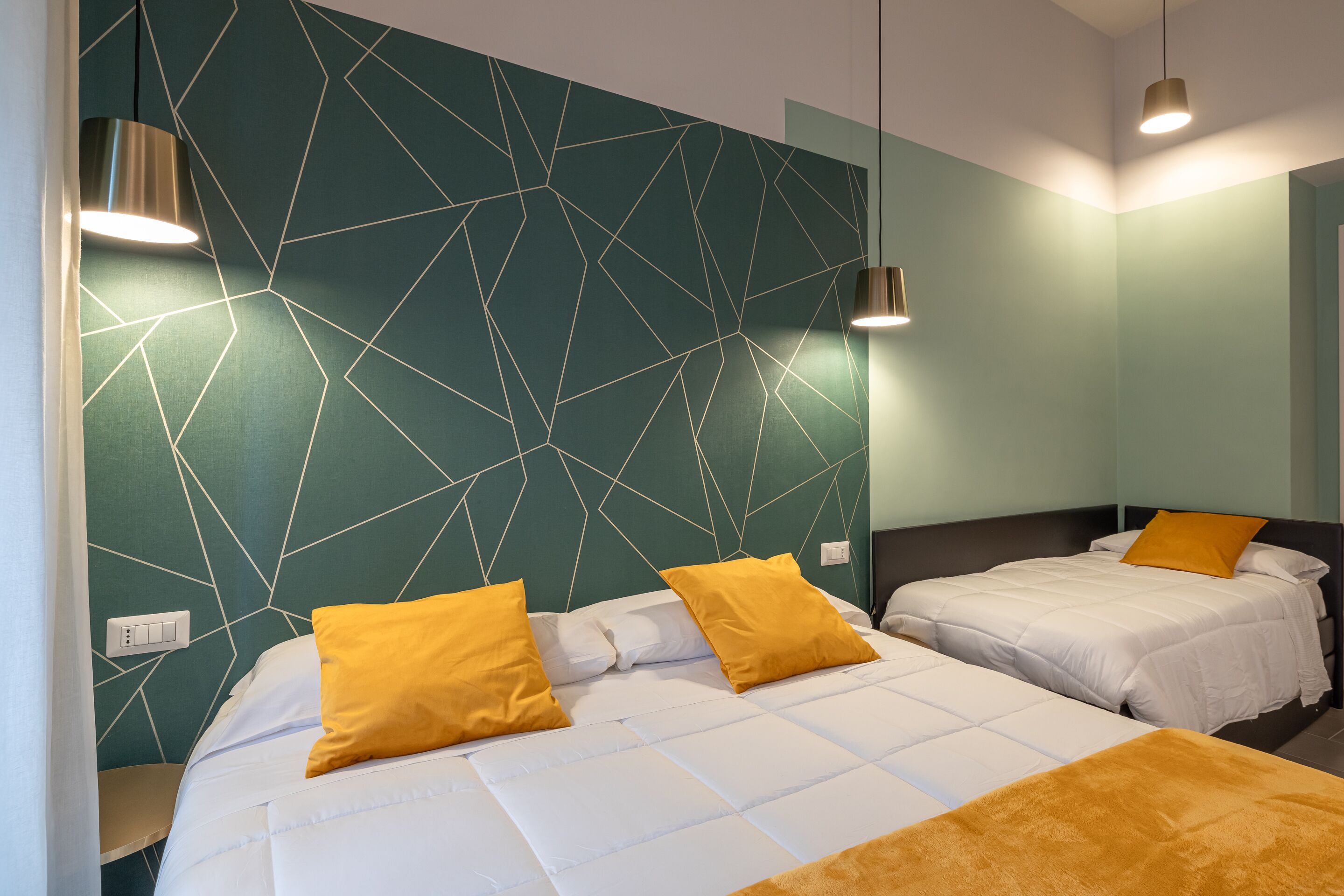 Habitación de hotel con dos camas, cada una con ropa blanca y almohadas amarillas. Las paredes tienen un diseño geométrico, y hay lámparas colgantes sobre las camas.