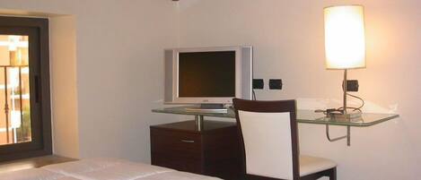 A bedroom with a bed, a desk with a computer, a chair, and a wall-mounted lamp.