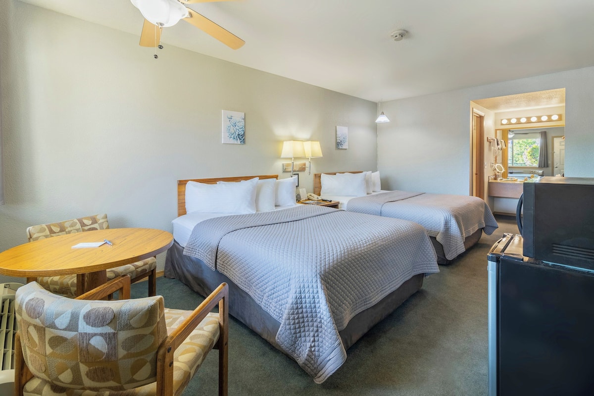 A hotel room with two beds, a dining table, chairs, a refrigerator, a ceiling fan, and a bathroom with a mirror and lights.