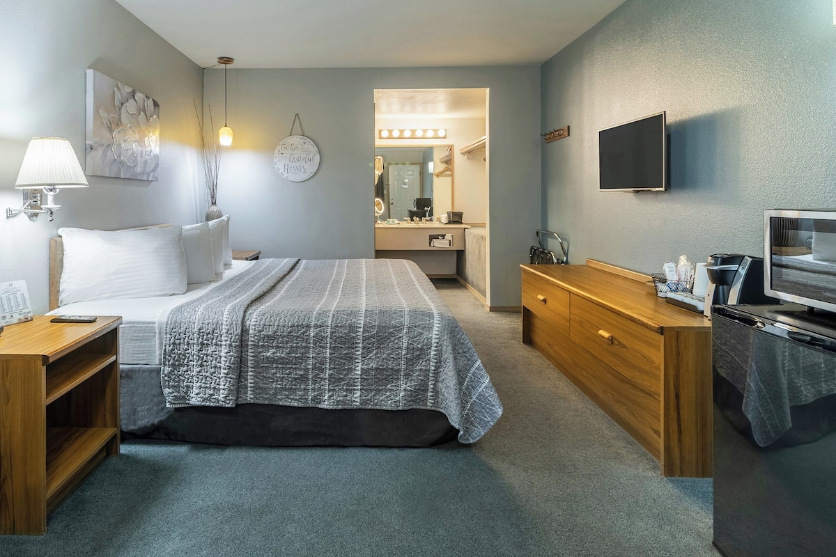A hotel room with a bed, bedside table, lamp, TV, desk, and a bathroom visible in the background.