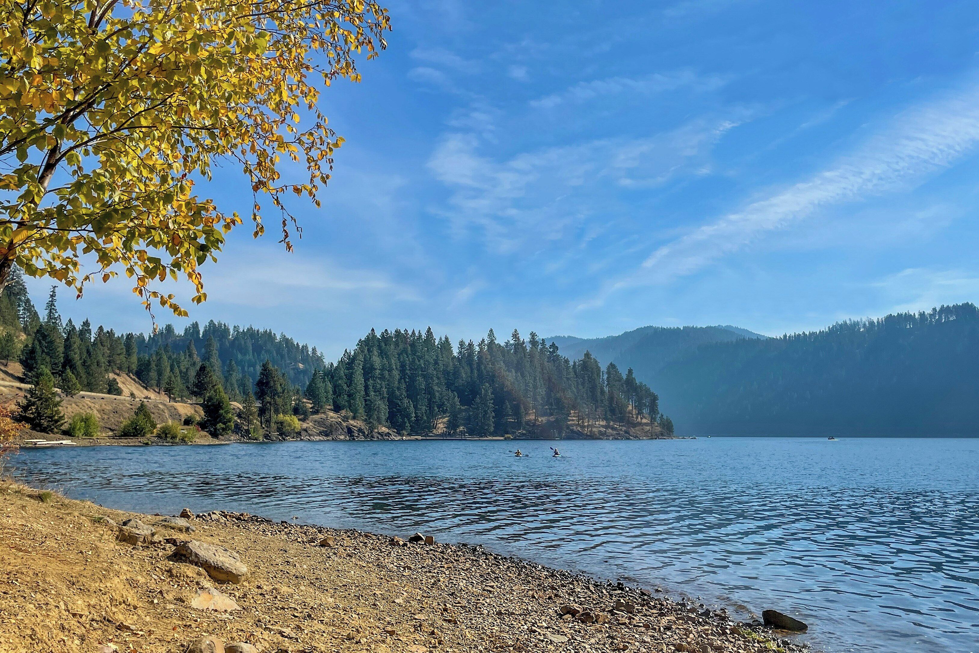A calm lake with kayaks, a rocky shoreline, and a forested mountain in the background.