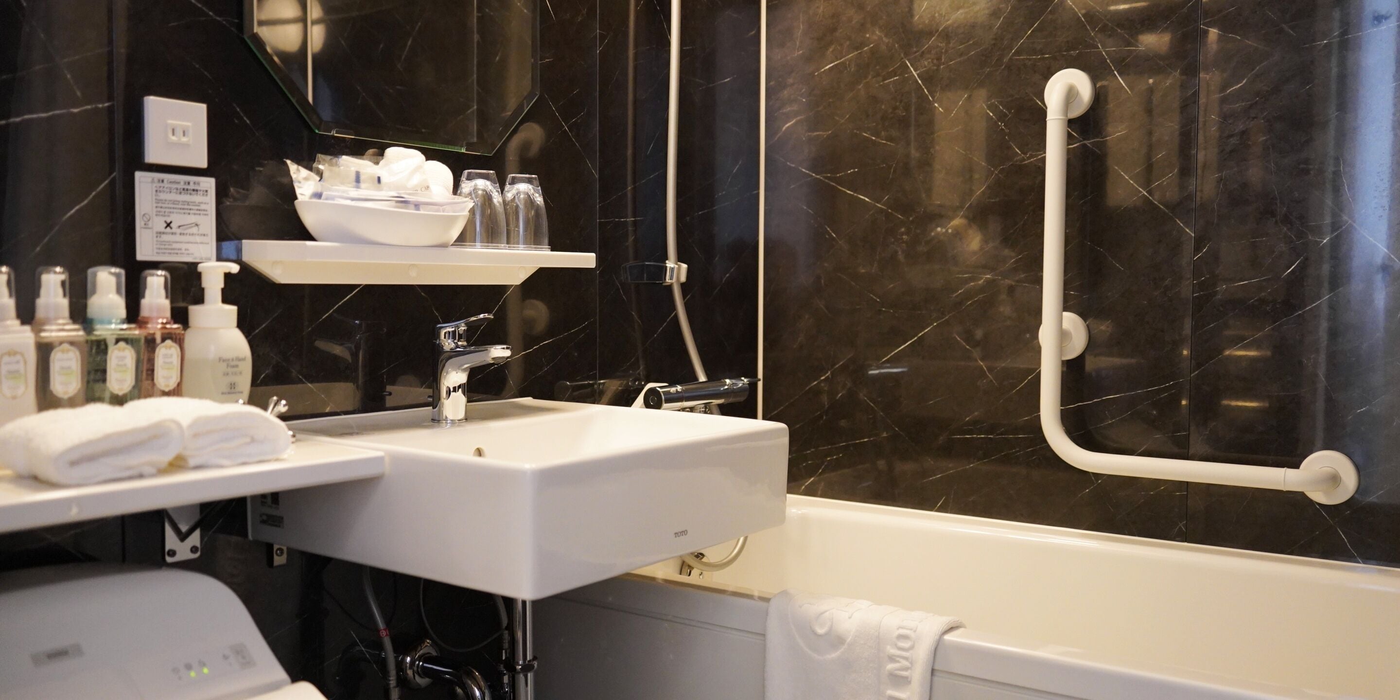 A bathroom with a white sink, a mirror, a shelf with toiletries, a bathtub, and a handrail.