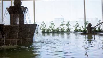 A water feature with a stone fountain in a pool, surrounded by greenery and a cityscape view.
