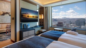A hotel room with a large bed, a desk with a flat-screen TV, a sofa, and a view of a cityscape and mountains.