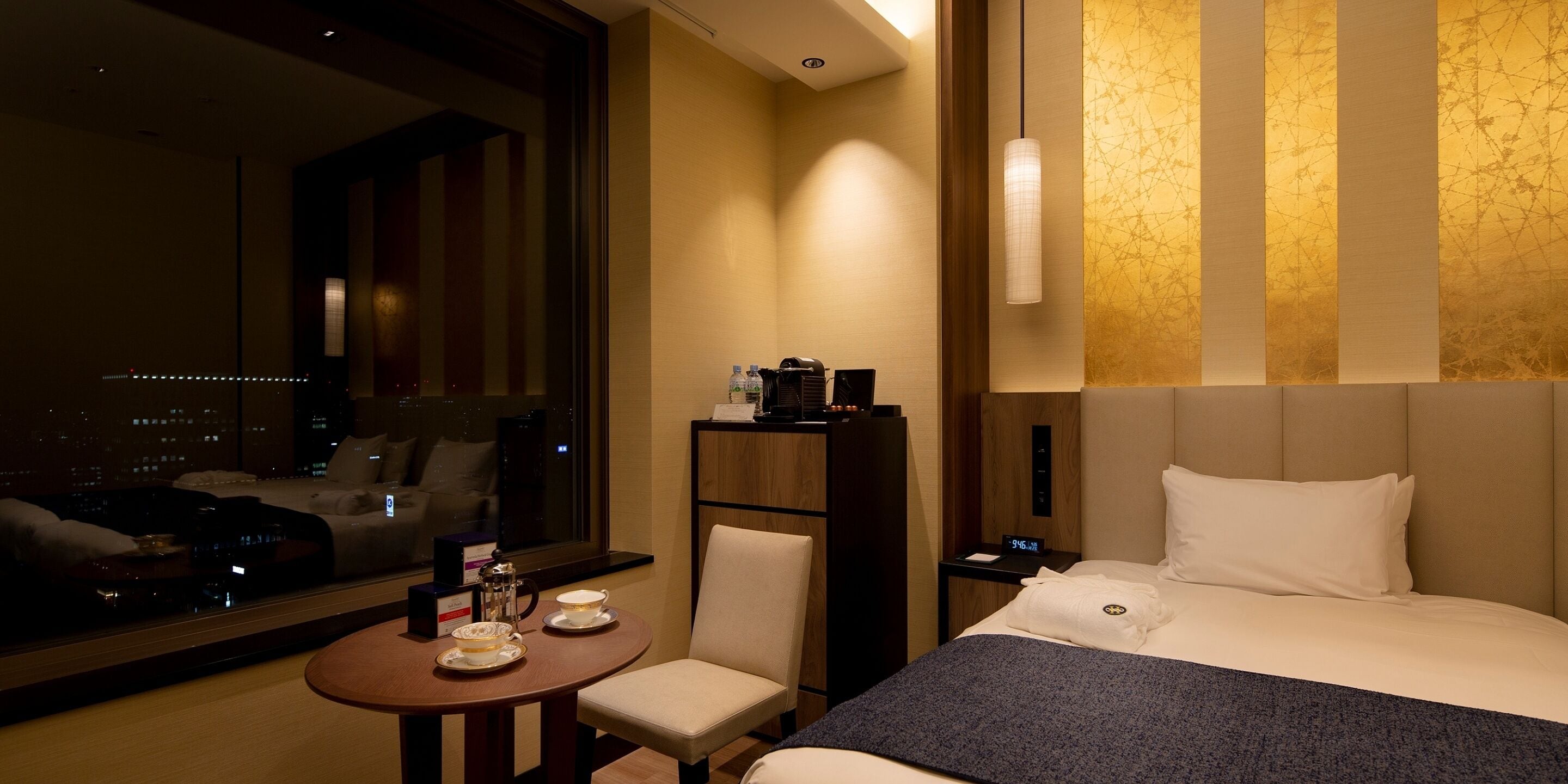 Ein Hotelzimmer mit einem großen Bett, einem Nachttisch mit Lampe, einem Sessel, einem kleinen Tisch mit Tee-Service und Blick auf die Stadt durch ein großes Fenster.