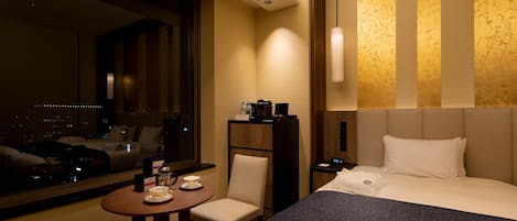 A hotel room with a large bed, a bedside table with a lamp, a chair, a small table with tea sets, and a view of the city through a large window.