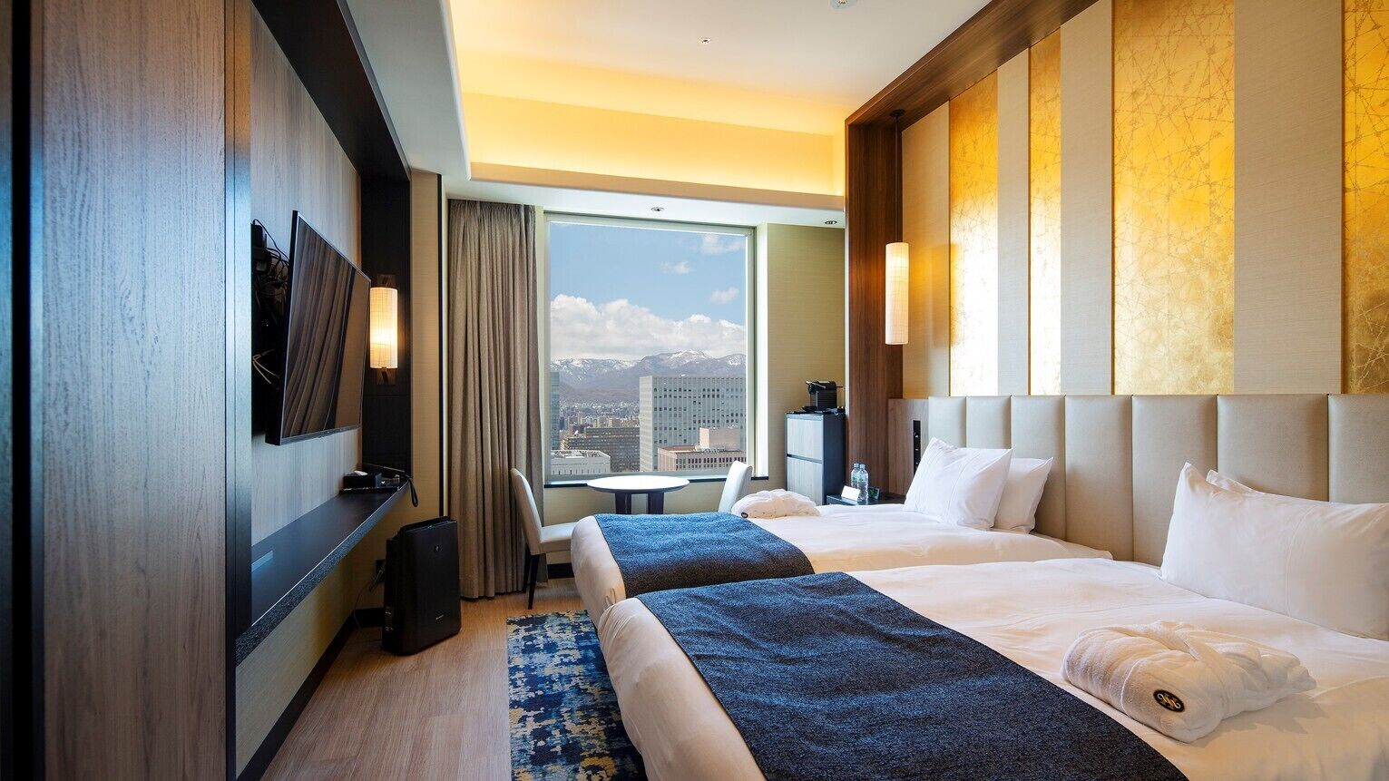 Ein Hotelzimmer mit zwei Betten, einem großen Fenster mit Blick auf die Stadt, einem kleinen Tisch und einem an der Wand befestigten Fernseher.