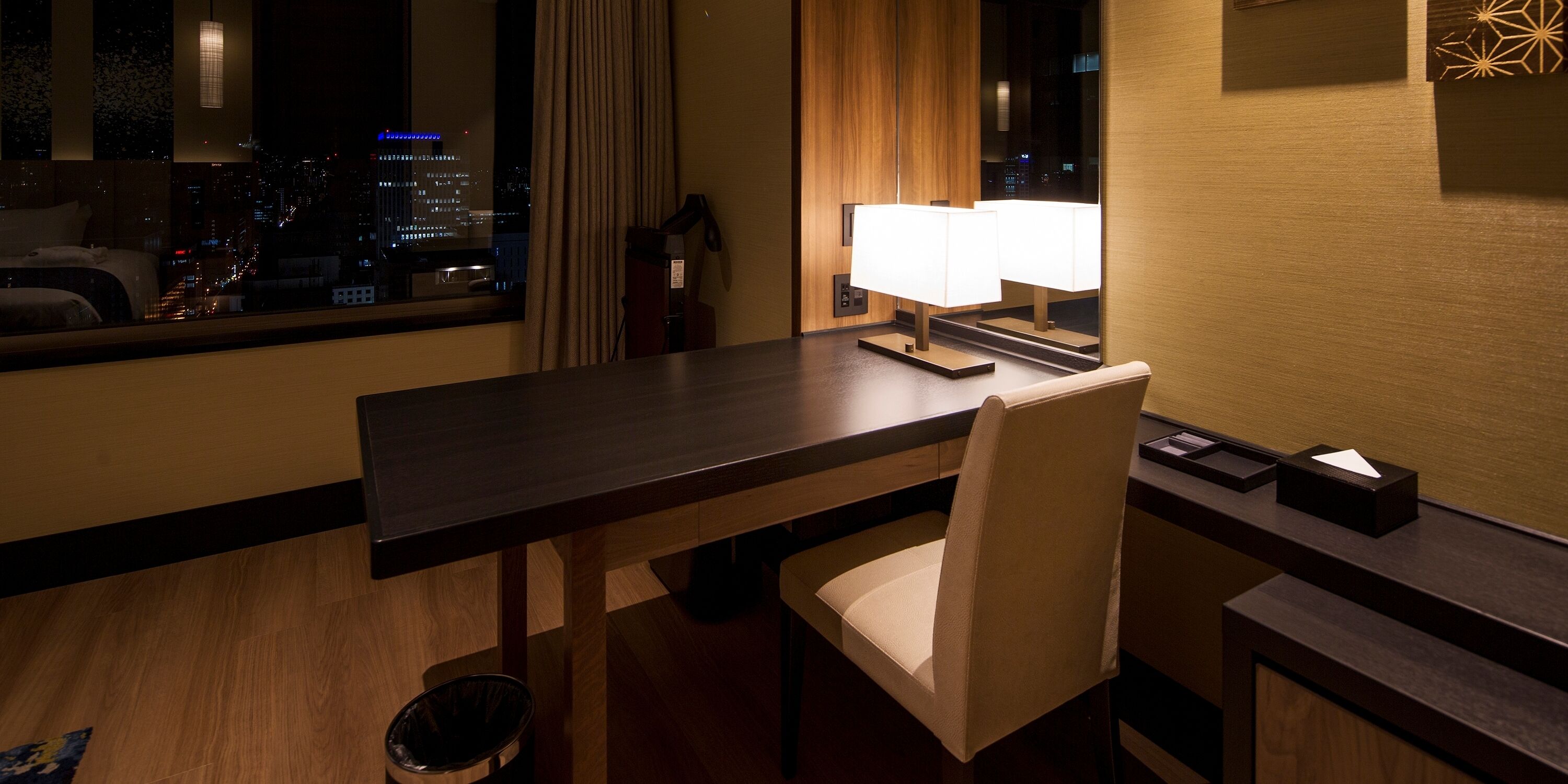 A hotel room with a city view, a wooden desk, a chair, a lamp, and a trash bin.