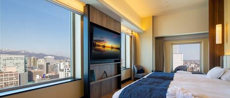A hotel room with a large bed, a TV mounted on the wall, and a city view through the windows.