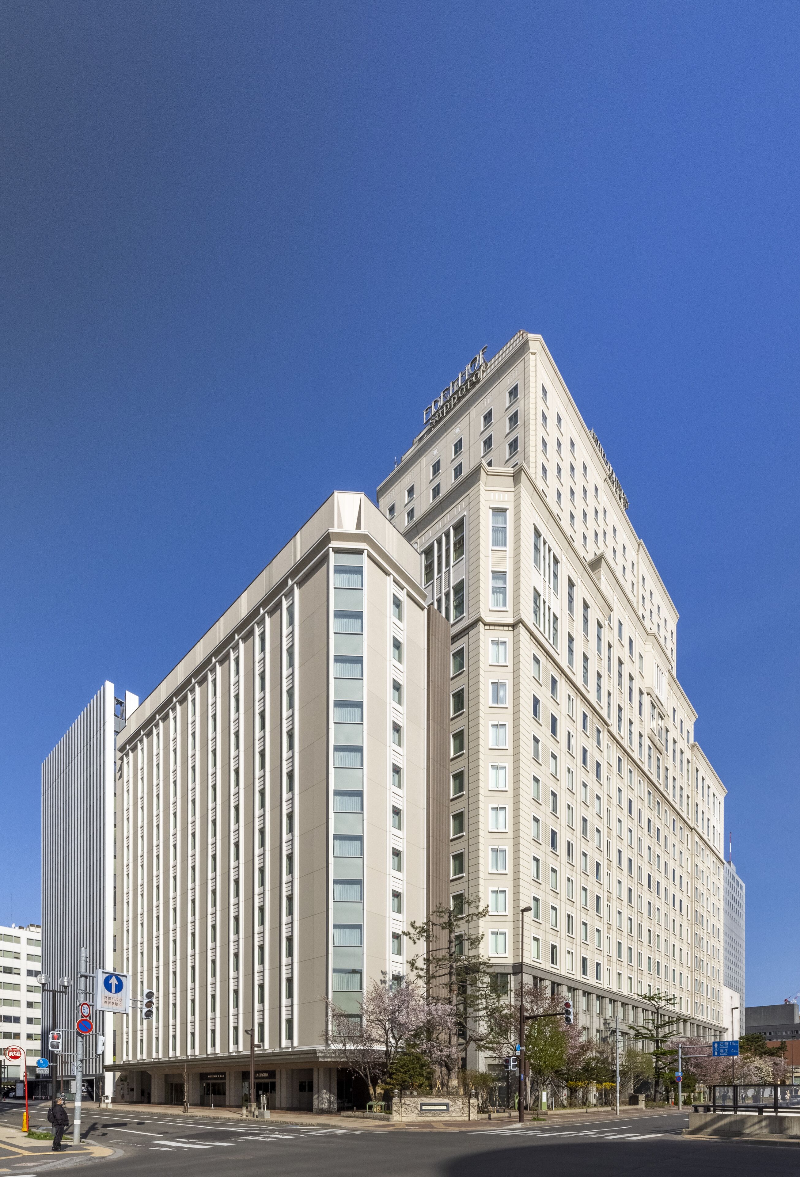 A large, multi-story hotel with a prominent corner entrance, a clear sky, and a few trees in front.